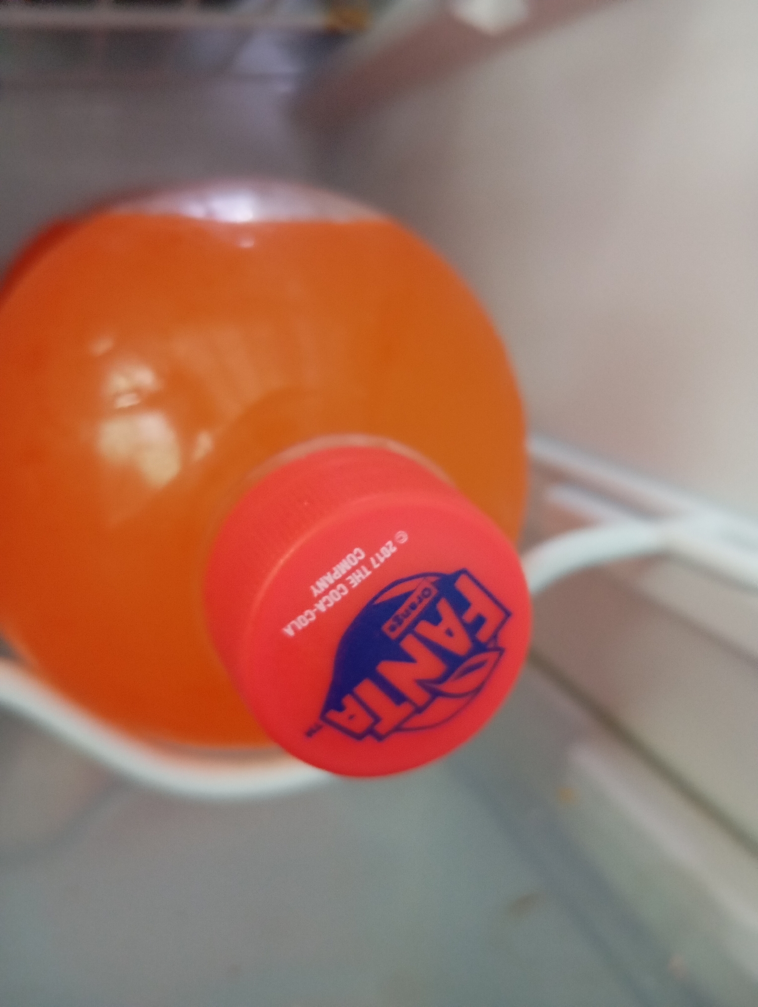 An orange Fanta bottle with a red cap is inside a refrigerator. The cap has the Fanta logo in blue and white, along with "2017 THE COCA-COLA COMPANY" printed in white text. The bottle is mostly in focus, while the background of the refrigerator shelf is slightly blurred.