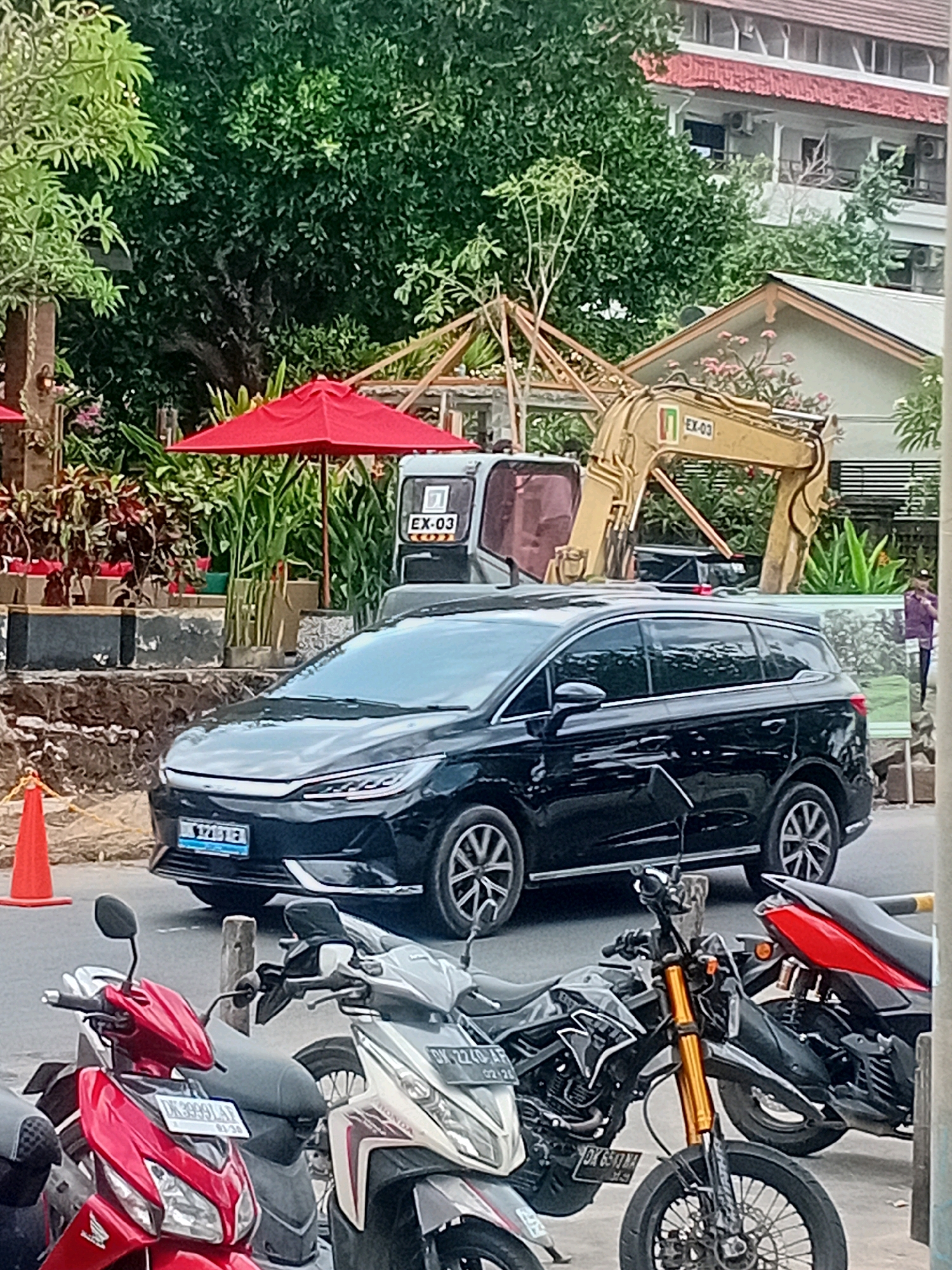 In Kuta, Indonesia, a black minivan is parked on the street, facing the camera. The minivan has a license plate that reads "DK 1712 EB". Several motorcycles are parked in front of the minivan. To the left of the minivan, there is a red traffic cone. In the background, a yellow excavator is visible, along with trees and buildings. The scene appears to be a street in a commercial or residential area, with construction ongoing.