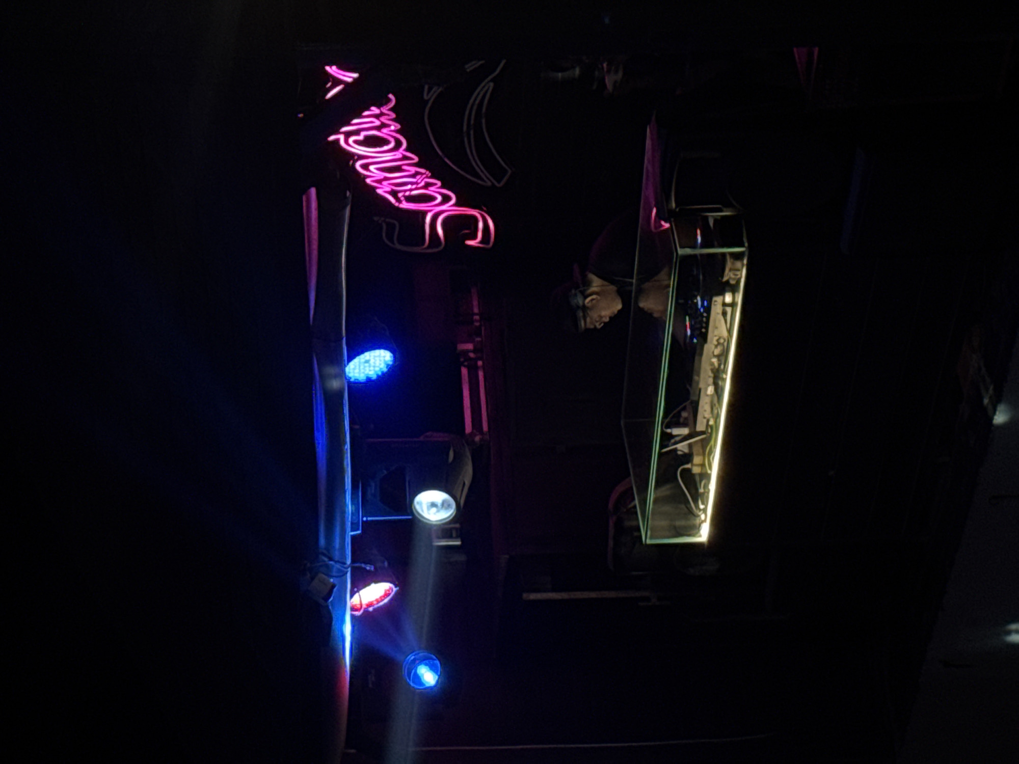 The image depicts a dimly lit interior, possibly a bar or nightclub, in Canggu, Indonesia. A neon sign in bright pink spelling "Smooth" is visible near a stage lighting rig with blue, red, and white spotlights. A glass box with a yellow LED strip contains electronic equipment. A person's reflection is faintly visible inside the glass box. The overall environment is dark, with limited illumination provided by the neon signs and stage lights.
