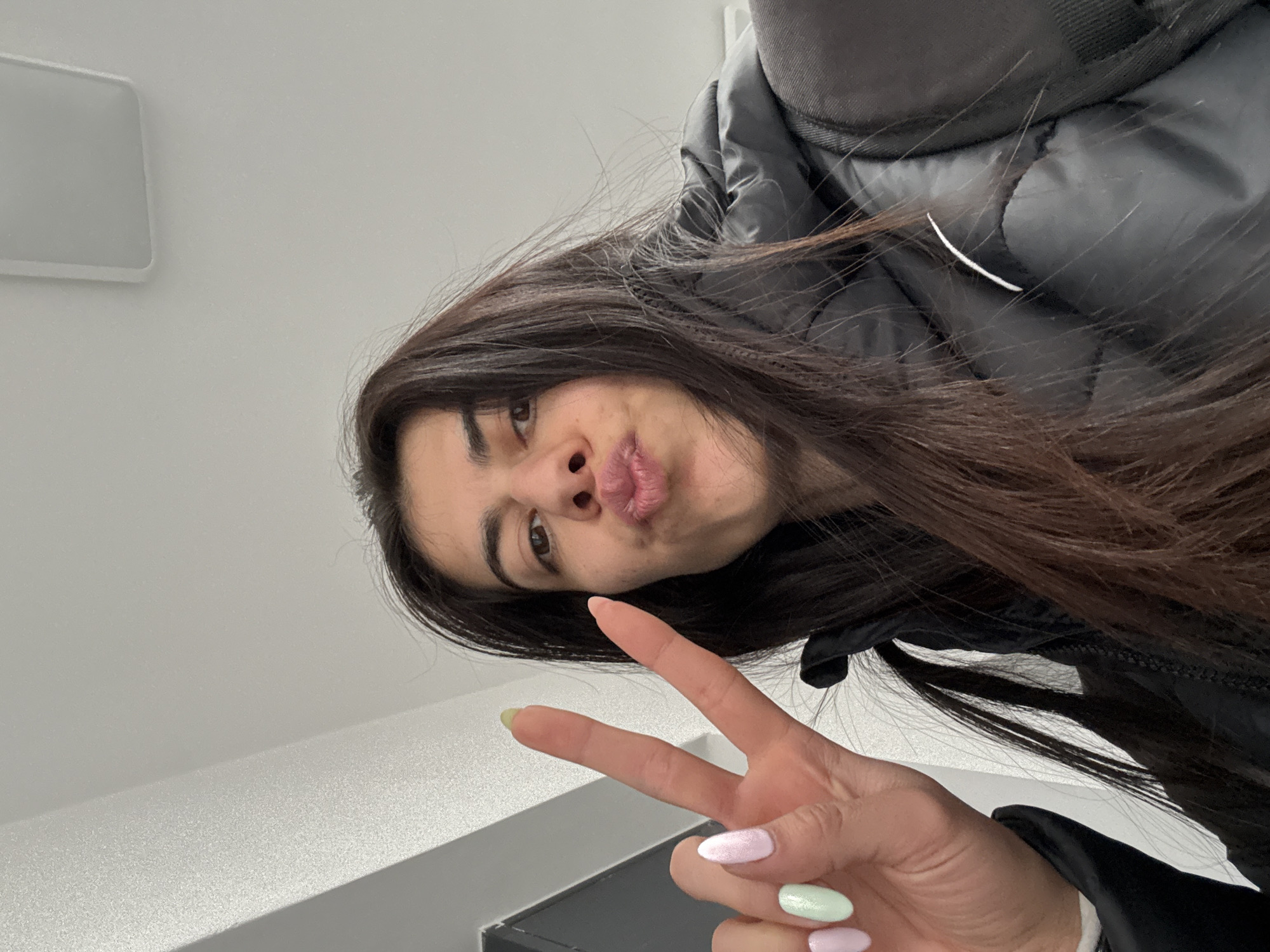 A young woman with long brown hair and a black puffer jacket is taking a selfie indoors. She is making a kissing face and holding up a peace sign with her hand, which has painted fingernails in pastel colors. The background is a white wall with a white rectangular light fixture and a grey surface. Location is indeterminate, but the city is Sofia, Bulgaria.