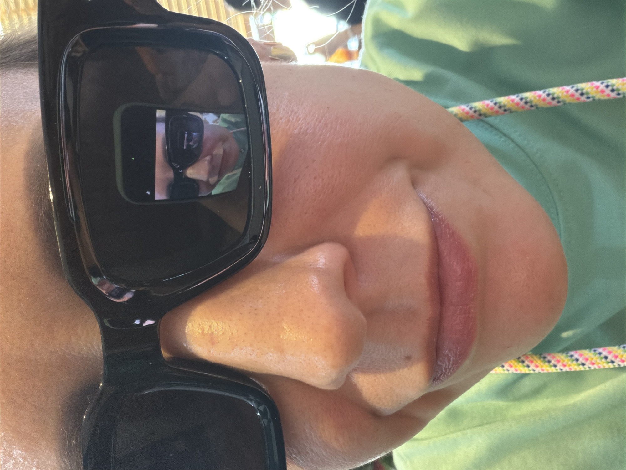 The image is a close-up, frontal shot of a person wearing large, black-framed sunglasses. The person's nose, lips, and chin are visible in the foreground. The sunglasses reflect a second image of the same person, also wearing sunglasses and holding up a phone to take the picture. The reflection shows the person's face and upper body, and the phone screen is also visible, displaying a green dot, likely indicating a camera recording or active status. The person appears to be outdoors, with a bright, somewhat blurred background that suggests natural light. The person is wearing a light green shirt, and a colorful braided lanyard is visible hanging down from the neck area of the shirt. No specific city or geographical features of South Quy Nhon Ward, Vietnam are discernible from the image.