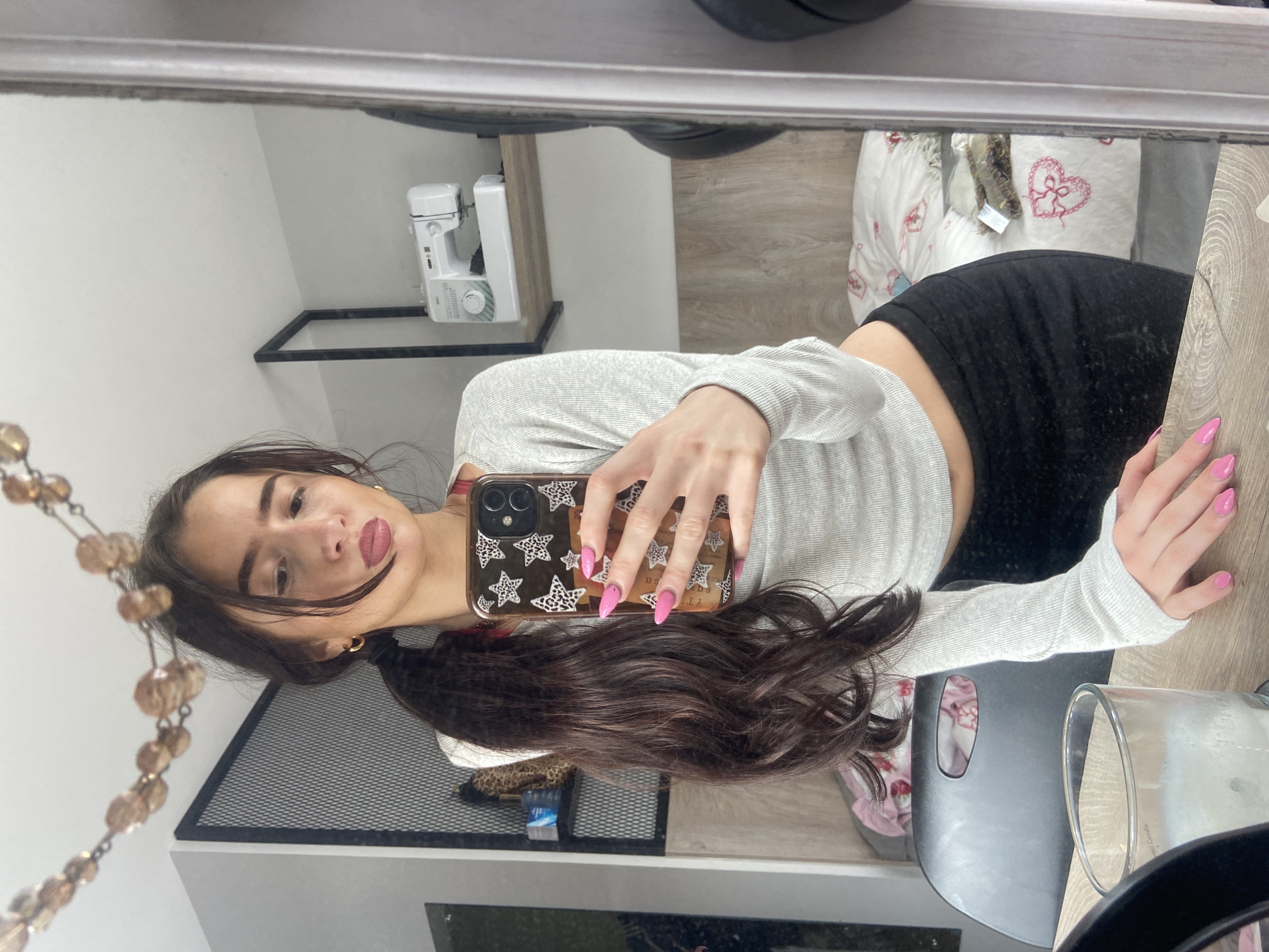 The image depicts a young woman taking a selfie in a mirror. She is wearing a long-sleeved grey top and black bottoms. Her long brown hair is styled in a ponytail. She holds a mobile phone with a star-patterned case in her right hand, her pink painted nails visible. The reflection shows a room with a sewing machine, a decorative black metal shelf, a heart-patterned pillow on a bed, and a large clear glass candle holder on a table. The background features elements typical of a domestic interior, suggesting an apartment or bedroom. There is no indication of the city of London.