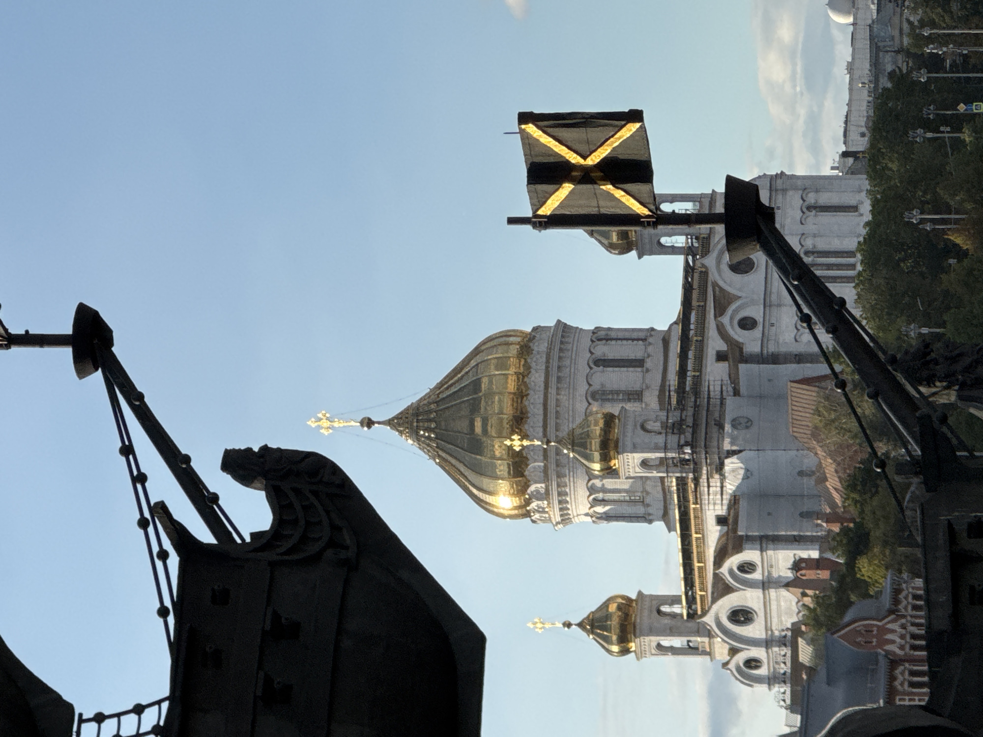The image shows a close-up view of the Christ the Savior Cathedral in Moscow, Russia. The cathedral, with its distinctive golden domes, is a prominent landmark in the city. In the foreground, the silhouette of a large, dark structure, possibly a statue or monument, partially obscures the view. A black flag with a yellow "X" pattern is mounted on a pole in front of the cathedral. The sky is clear blue, with a few wispy clouds. Trees and other buildings can be seen in the background, suggesting an urban setting. The lighting indicates it is daytime, with the sun casting a warm glow on the cathedral's domes.
