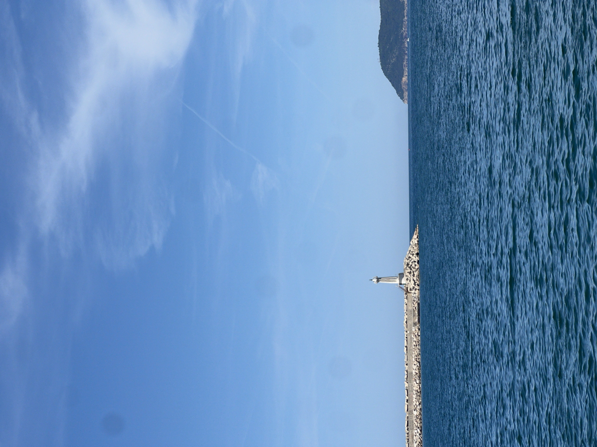 The image displays a coastal scene, rotated 90 degrees clockwise. When viewed correctly, it shows a wide expanse of bright blue sky dominating the upper half, meeting a deep blue sea at the horizon, which extends across the lower half.
In the mid-ground, a white lighthouse stands prominently at the end of a long, gray breakwater constructed from rocks, stretching horizontally across the water. The sea exhibits a texture of numerous small, parallel ripples, suggesting calm but moving water. On the distant horizon, a dark, rocky, and possibly forested mountain or hill is visible on the far right, partially obscured by the sea and sky.
The sky is a clear, vibrant blue, with scattered white, wispy clouds (likely cirrus) and a few faint contrails from aircraft, indicating a high-altitude atmospheric presence. The lighting suggests it is daytime, likely mid-morning or early afternoon, with bright sunshine.
There are no people or specific activities depicted in the image. No visible text is present. The overall setting is serene and open, capturing a tranquil coastal environment in Bar, Montenegro, with a focus on natural elements and a key navigational structure.