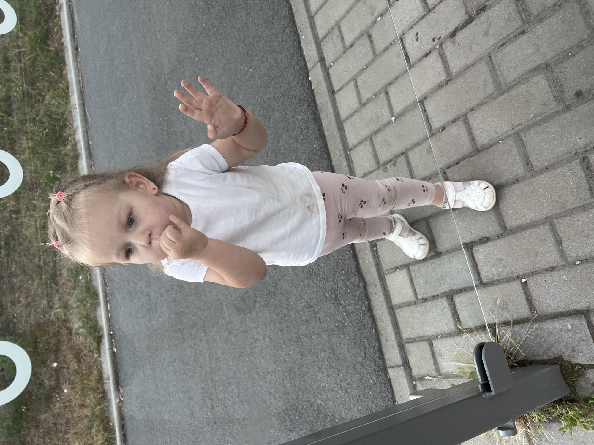 A young girl with blonde hair in two pigtails and wearing a white t-shirt and pink leggings with a cherry pattern is standing on a paved area next to a road. She is putting her fingers in her mouth and looking to her left. A red bracelet is visible on her right wrist. The girl is standing in Pilsen, Czechia.