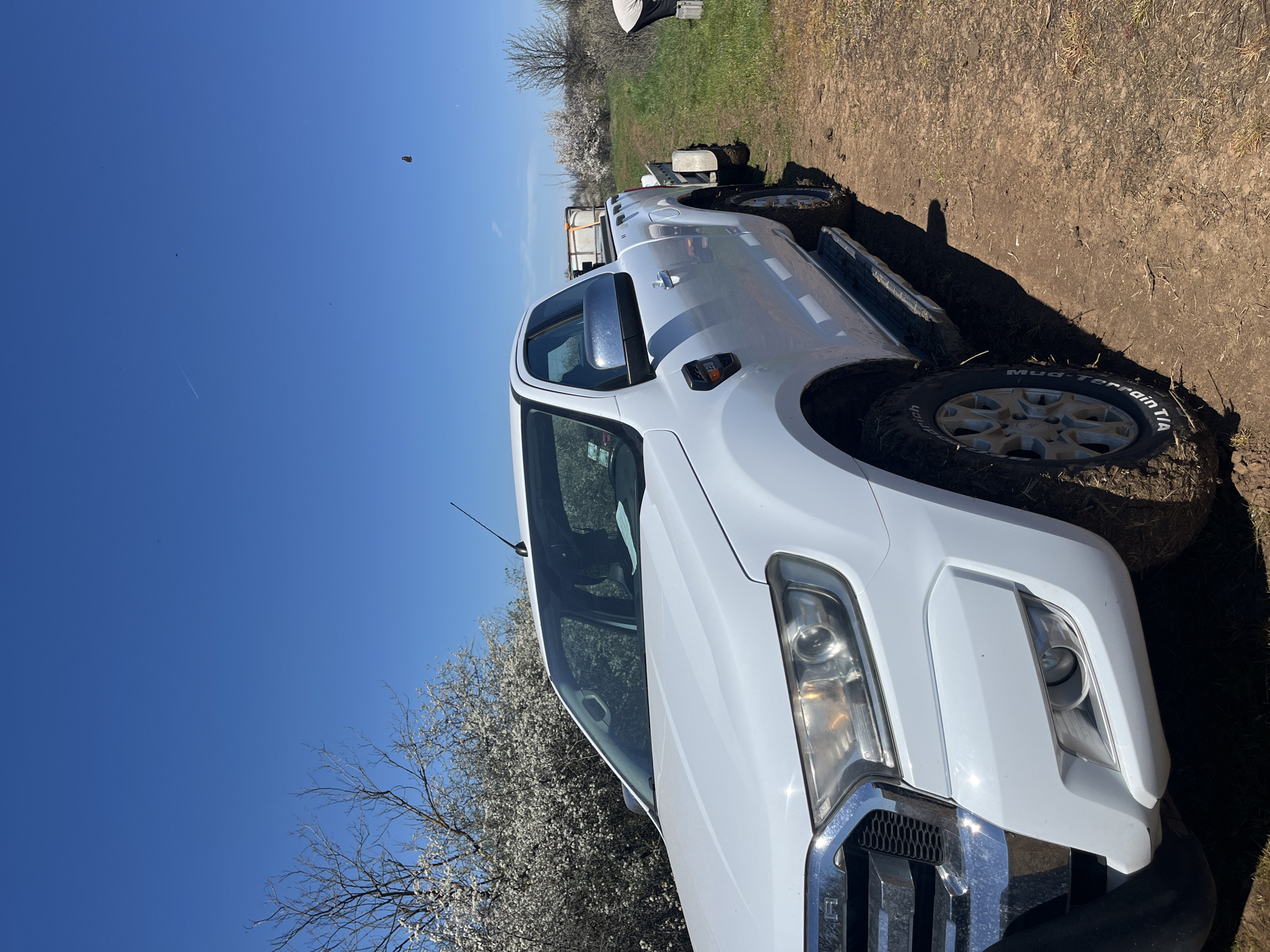 The image shows a white pickup truck parked on a dirt and grassy area, possibly a field or rural location in the city of Unknown, Unknown. The truck is angled towards the viewer, showcasing its front passenger side. A trailer is hitched to the back of the truck. A tree with white blossoms is visible on the left, and the sky is clear blue. The front tire is close to the dirt and the tire reads "Mud-Terrain T/A"