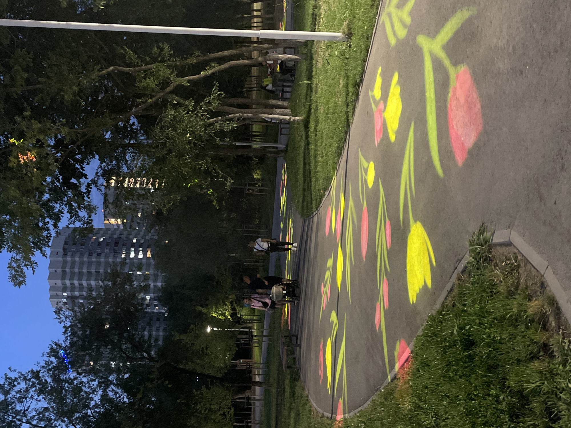 This is an evening, outdoor, medium-shot photograph of a paved park pathway in Almaty, Kazakhstan. The pathway is illuminated with projected images of large, stylized tulips in pink and yellow, with green stems and leaves. The pathway curves from the lower left to the upper right of the frame. On the left side of the pathway is a strip of mown grass. In the background, behind trees, are tall, modern apartment buildings with many lit windows. A white lamppost stands on the right side of the path. Three people are walking on the path: a woman with a pink bag, a man, and another person pushing a stroller. Further down the path, two individuals are sitting at a picnic table. The sky is a deep blue, indicating dusk.