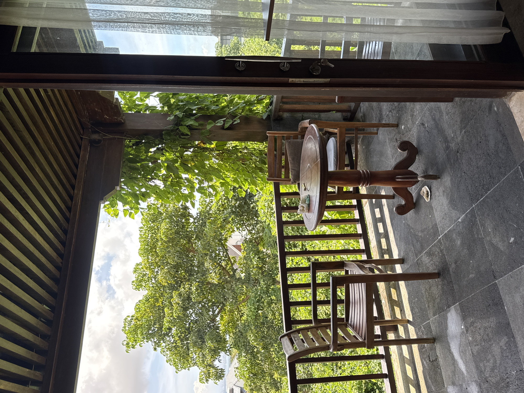 The image depicts an outdoor patio in Ungasan, Indonesia. The patio features a round wooden table and chairs, set on a grey tiled surface. A wooden railing surrounds the patio, with lush green trees visible beyond. The view is partially framed by a doorway with sheer white curtains. The sky, with scattered clouds, is visible through the foliage.