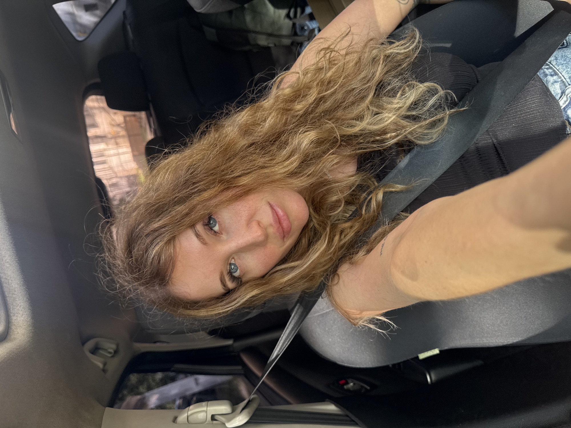 The media file features a young woman with long, wavy blonde hair in a car. She is looking directly at the camera, with a slight smile and blue eyes. The woman is wearing a black top and denim jeans. A black seatbelt is visible across her chest, indicating she is likely the driver or a passenger. The interior of the car is dark, with glimpses of the outside through the windows. The scene appears to be set during the daytime, with natural light filtering into the car. The overall mood is casual and relaxed. No visible text is present in the image. Based on the provided location context, the scene is likely taking place in Chaikovsky, Russia.