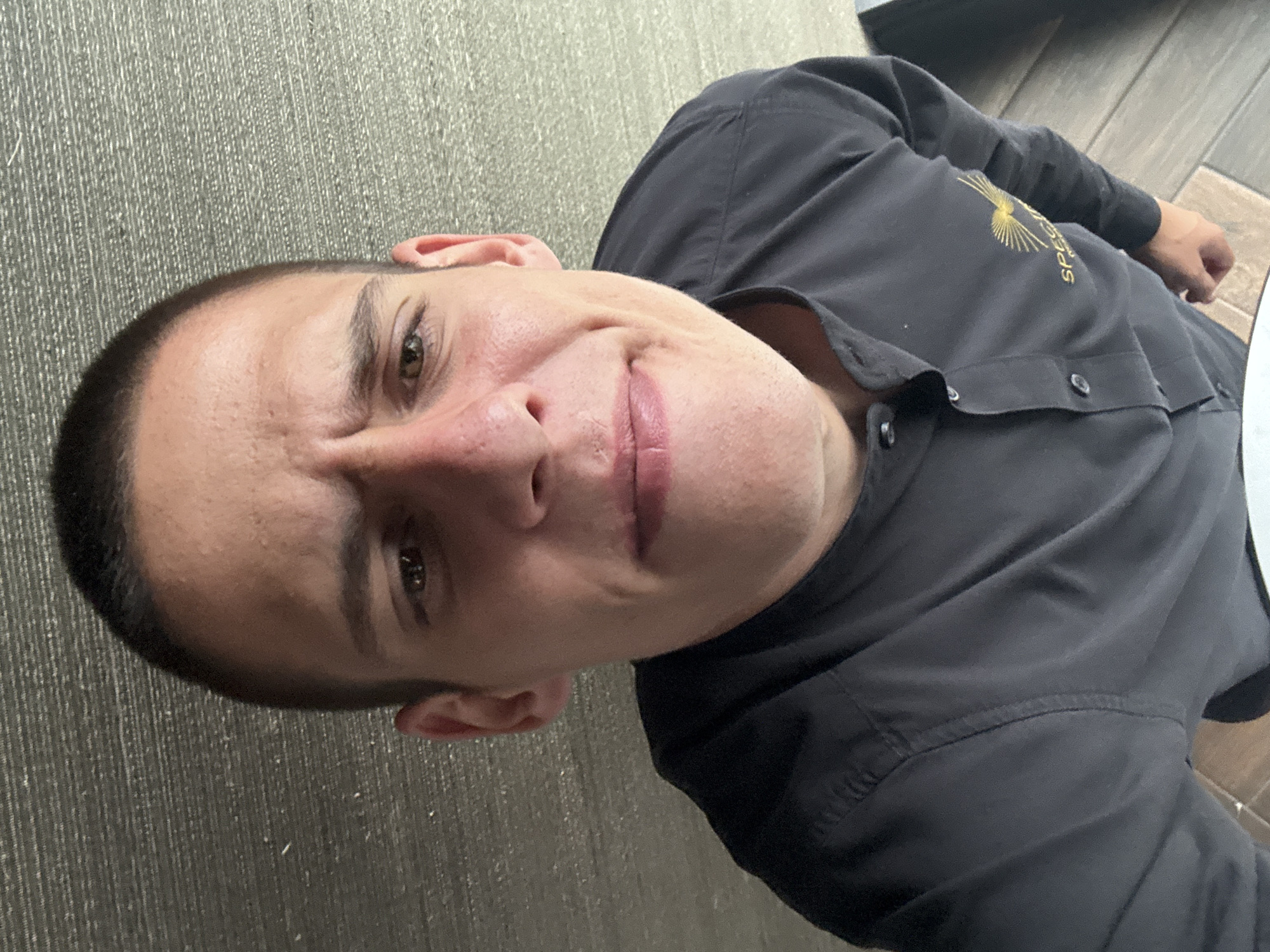 A close-up, high-angle selfie shows a young man with short, dark hair and light brown eyes. He is wearing a black collared shirt with buttons down the front. A gold logo with radiating lines is visible on the left side of his chest. His facial expression is one of mild consternation, with his eyebrows furrowed and his mouth slightly pursed. He appears to be in an indoor setting, with a textured grey wall behind him and a wooden floor beneath. The image's timestamp indicates it was taken on March 29, 2023, at 09:37:02. The location data associated with the image places it in Sofia, Bulgaria.