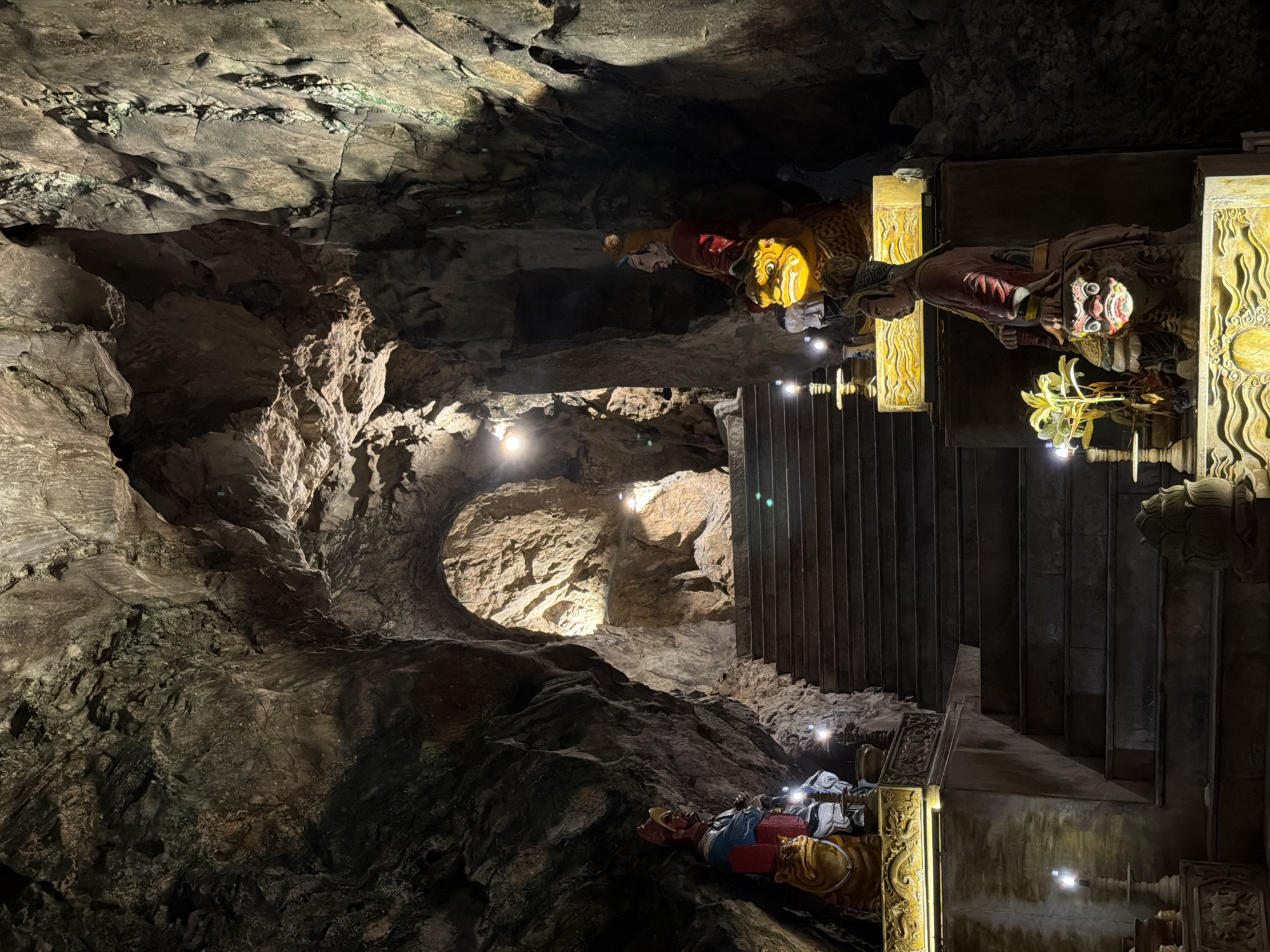 The image depicts the interior of the Marble Mountains cave complex in Đà Nẵng, Vietnam. The cave's rock formations are visible, illuminated by artificial lighting. A set of stairs ascends into the cave, flanked by ornate statues of deities or mythical figures, positioned on either side of the stairway. These statues are decorated in gold and red colors, with intricate carvings on their bases and surrounding architectural elements. Some people are visible near a statue in the lower portion of the cave.