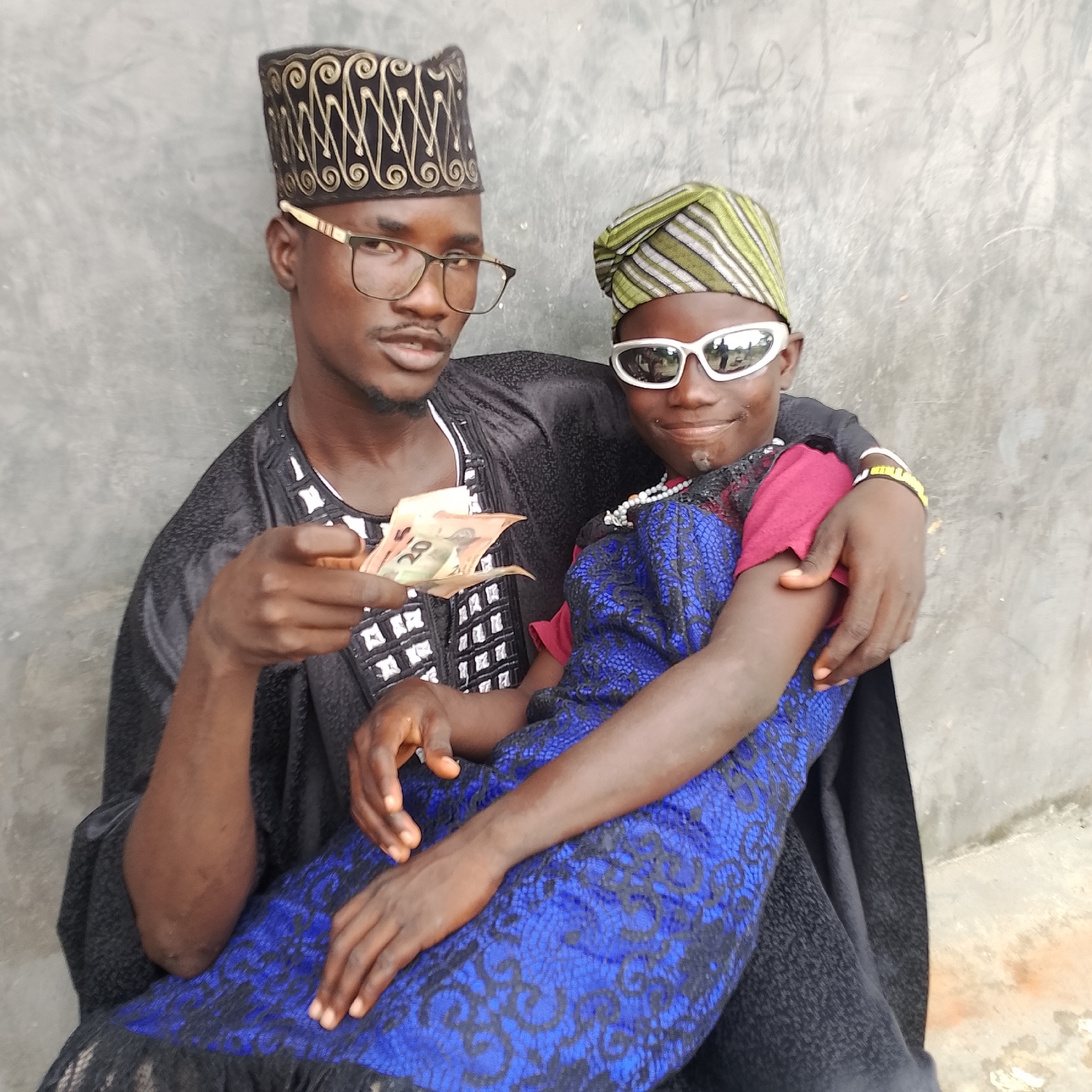 Two individuals, likely male, are pictured in what appears to be an outdoor urban setting in Ado, Nigeria. The person on the left, wearing a black embroidered cap and glasses, is holding out a few Nigerian Naira banknotes towards the camera. The second individual, seated on the left person's lap or close by, wears a green and white striped cap and white sunglasses, with their arm draped around the other person's shoulders. Both are dressed in traditional African attire. The background consists of a rough, grey concrete wall, typical of many urban environments in Nigeria. The lighting suggests it is daytime, with the sun casting shadows. The overall impression is a casual, candid photograph taken by one of the individuals or a third party.
