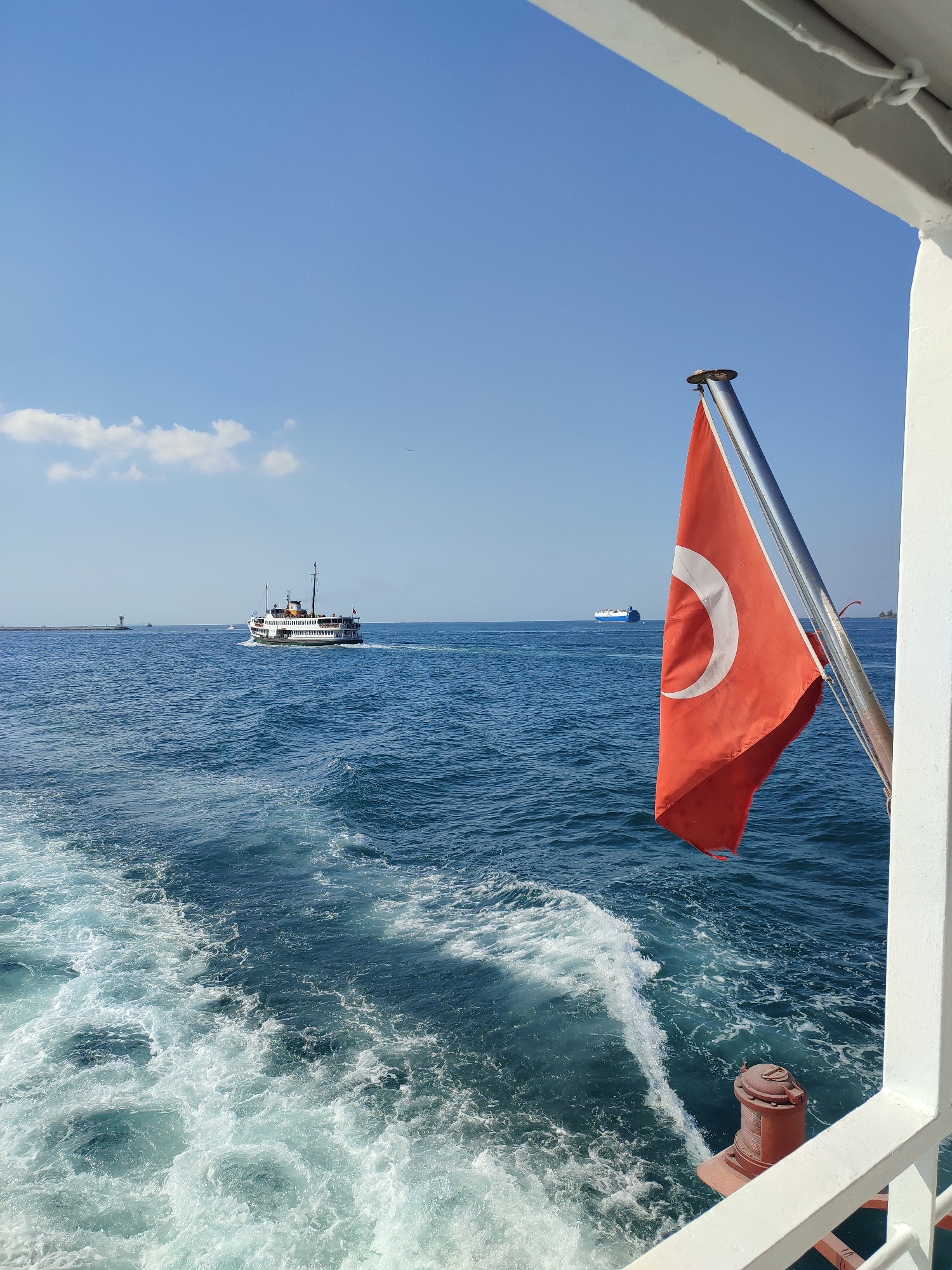 The image depicts a view from a moving ferry in Istanbul, Turkey. In the foreground, a Turkish flag with a red background, white crescent moon, and star waves from a metal flagpole attached to the ferry's white railing. The ferry is moving through a body of water, leaving a white foamy wake. In the mid-ground, a white passenger ferry with multiple decks and black accents travels in the distance. To the left, a lighthouse or navigational marker stands on a breakwater. Further out in the distance, a large blue and white cargo ship is visible on the horizon. The sky above is clear blue with a few wispy white clouds. The overall scene conveys a sense of maritime travel and the iconic seascape of Istanbul.