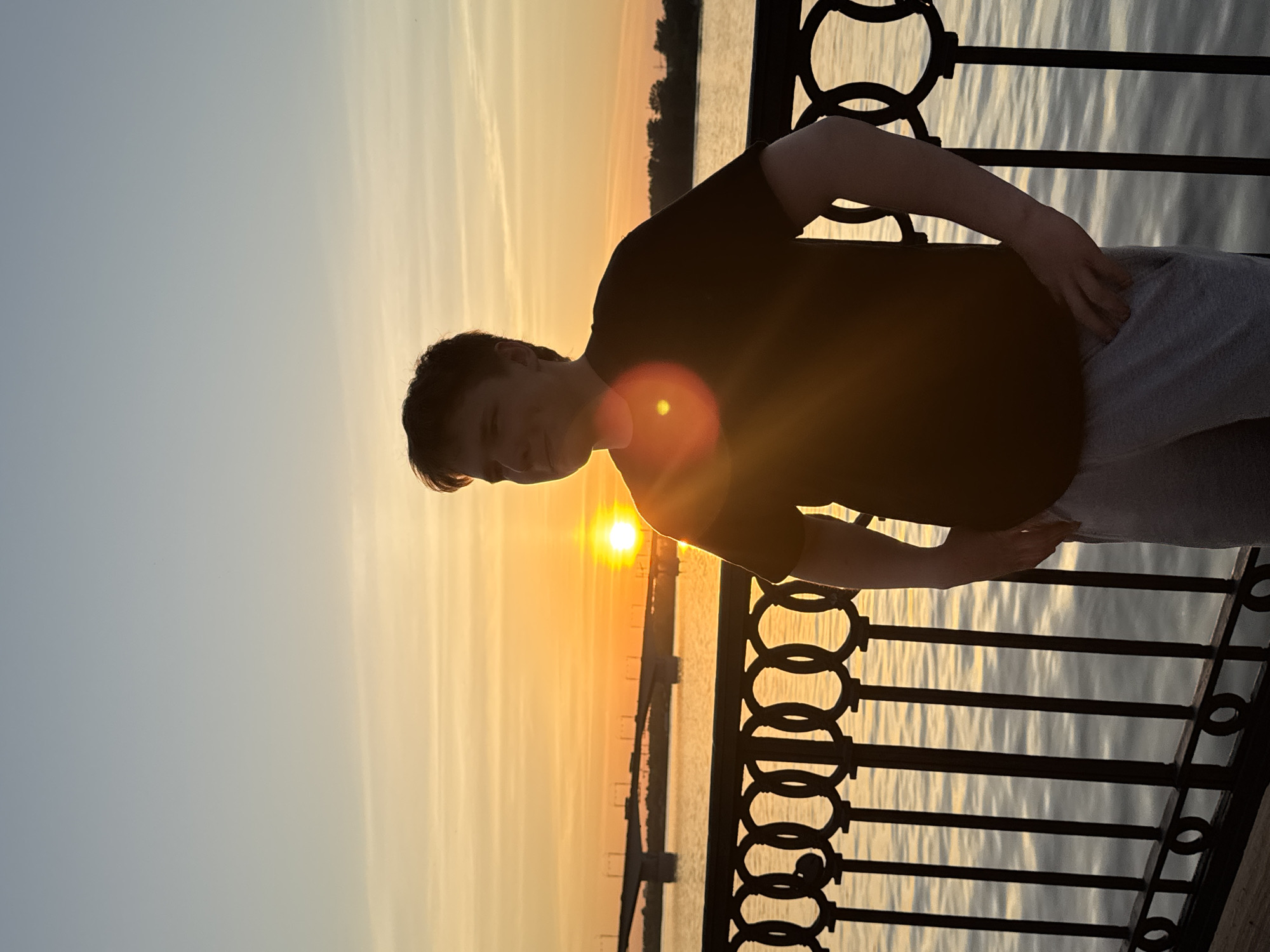 A young man is posing for a photo on a balcony or observation point in Perm, Russia, during a sunset. The sun is setting over the Kama River. He is wearing a black t-shirt and gray pants. He is standing next to a black metal railing.