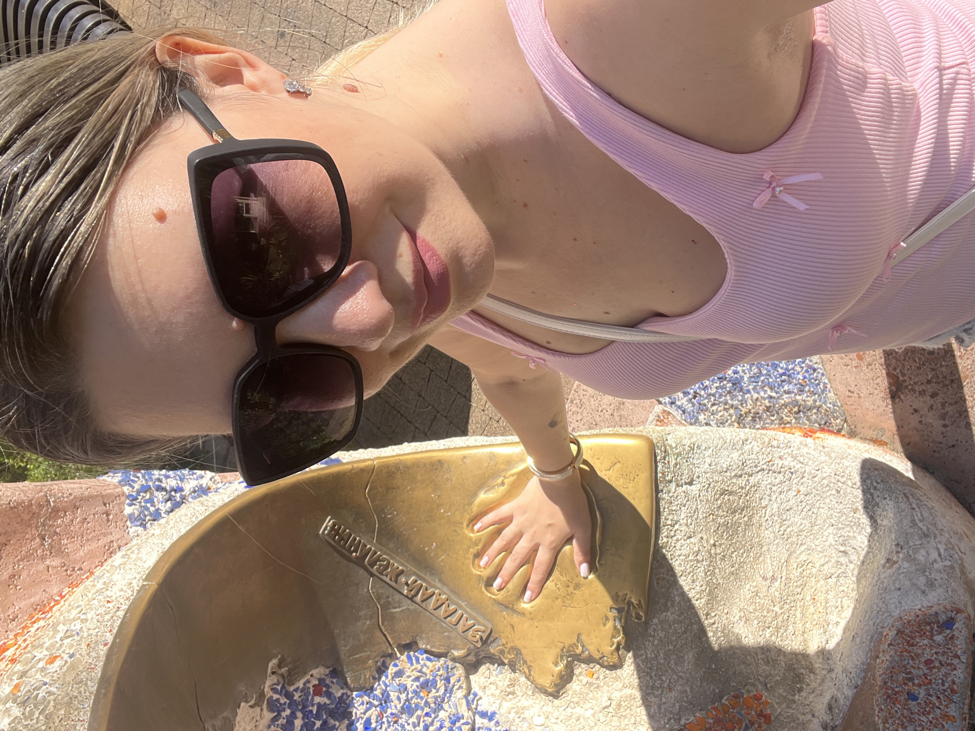 A young woman with blonde hair and sunglasses is lying down outdoors, with her head tilted back. She is wearing a pink ribbed tank top with small bow embellishments. Her right hand, adorned with a bracelet, is resting on a bronze plaque that appears to be embedded in a mosaic-covered structure. The plaque shows a handprint and some text. The scene suggests it is daytime, with bright sunlight casting shadows. The background includes textured surfaces and a glimpse of greenery, hinting at an outdoor park or public space. The overall mood appears relaxed and sunny. The visible text on the plaque is in Cyrillic, and appears to read "ЗОЛОТОЙ КОЛОС" which translates to "GOLDEN EAR OF WHEAT".