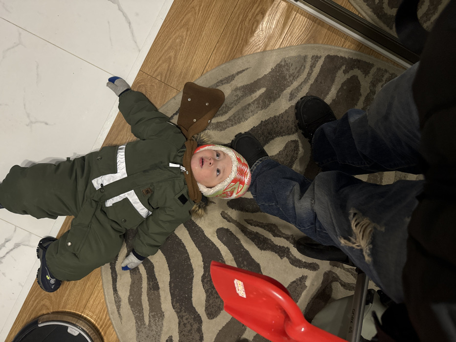 A young child in a green snowsuit and colorful hat lies on a zebra-striped rug. A brown floppy hat is near the child's head. Adjacent to the rug are a hardwood floor and white tiled floor. Nearby is a red toy shovel. A person wearing blue jeans and black shoes stands partly on the rug, partially obscuring the scene. A robotic vacuum cleaner is visible in the corner.