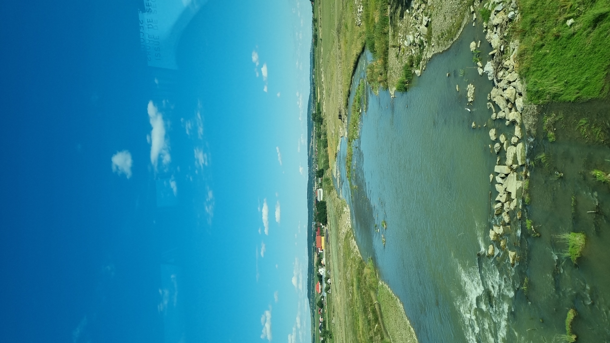 This is a landscape photograph taken from an elevated vantage point, looking down at a river flowing through a rural area. The sky is a vibrant blue with scattered white clouds. In the distance, a small town with red-roofed buildings is visible nestled amongst rolling green hills. The river itself is wide and somewhat shallow, with a rocky bottom visible in places. The banks of the river are lined with green grass and vegetation, with some areas of rocky terrain. A small cluster of white stones are arranged in the river, creating a slight ripple effect. The overall impression is one of a peaceful, rural setting on a clear, sunny day. The location is identified as Milișăuți, Romania.