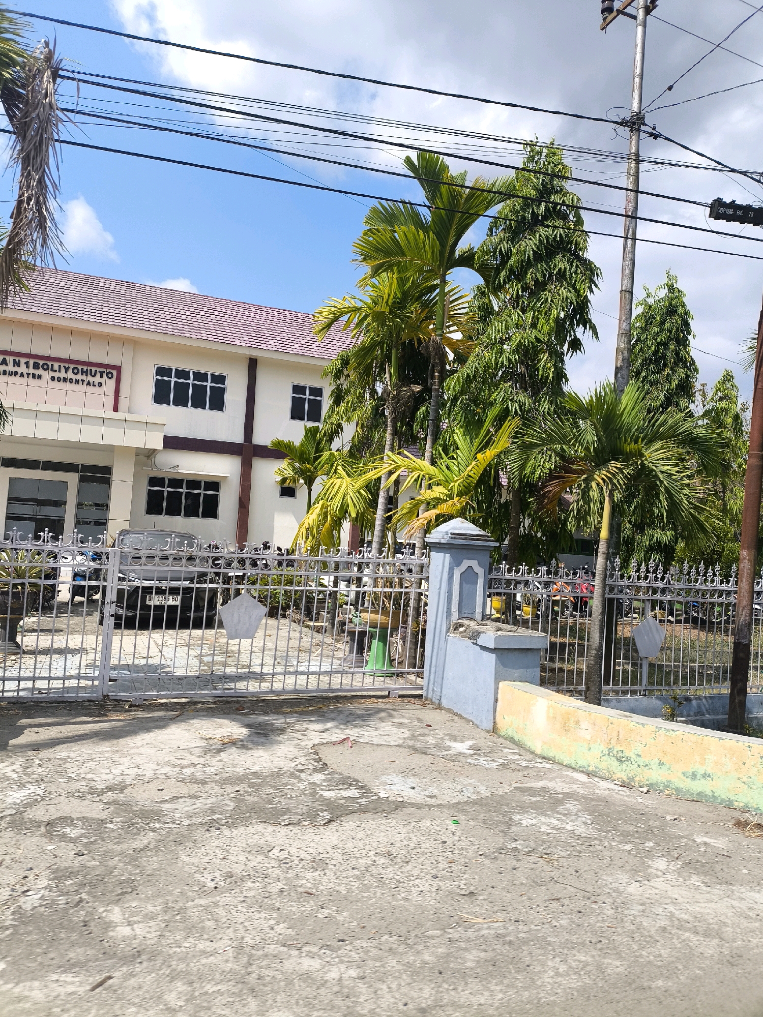 The image shows the exterior of a beige building with a red tiled roof, identified by text on the building as "MAN 1 BOLIYOHUTO KABUPATEN GORONTALO". The building has multiple windows with dark frames. In front of the building is a silver metal gate. Beyond the gate, a black car with a license plate that reads "DI 1183 BO" is parked. Several motorcycles are also visible behind the gate. Lush green trees, including palm trees, are present in the foreground and background, some of which are dry and brown. A utility pole with wires runs vertically through the right side of the image. A blue and gray concrete pillar stands near the gate. The ground in the foreground is a cracked concrete surface, with some patches of discoloration and scattered leaves. The sky above is blue with scattered white clouds. The scene appears to be a sunny day in Sido Mulyo, Indonesia.