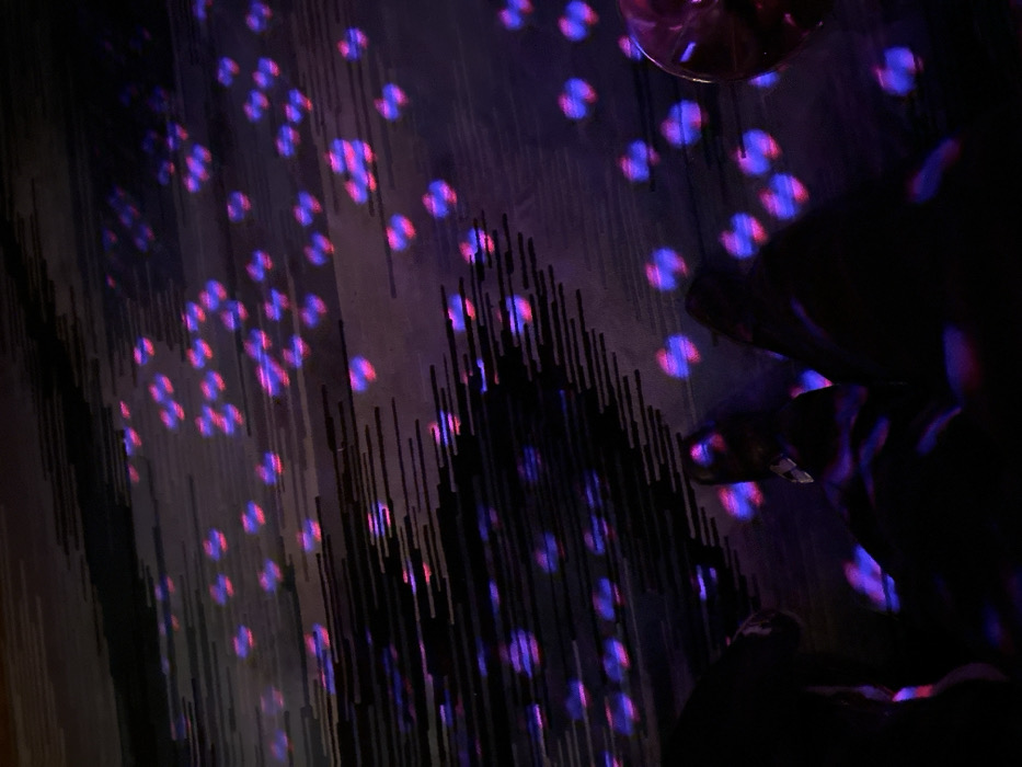 A dimly lit room features an abstract carpet pattern of vertical lines and scattered luminous spots in shades of purple and pink. A person's lower body and shoes are silhouetted against the light on the right side of the image, partially obscuring the carpet. A clear glass, likely a wine glass, is positioned in the upper right corner. The lighting creates a dynamic interplay of shadows and illumination.