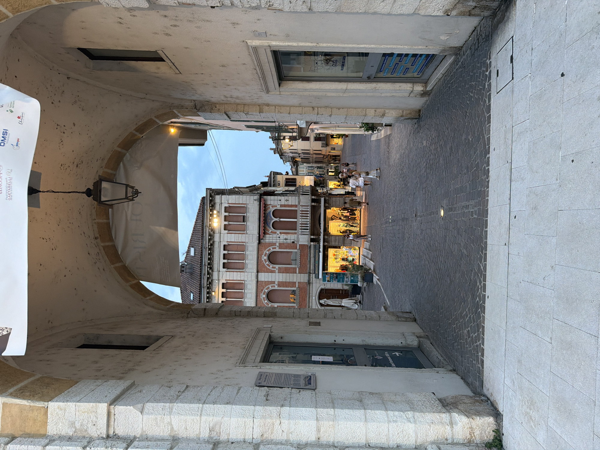 The image captures a view through an ancient stone archway leading into a cobblestone street in Salò, Italy. The archway, constructed of rough-hewn, light-colored stone blocks, frames a street scene bathed in the soft light of dusk. Above the arch, a banner is partially visible. A vintage lantern hangs from the ceiling of the arch.

The street beyond the archway is paved with dark cobblestones. On the left side of the street, a building with a red brick facade, featuring arched windows and decorative balconies, is prominent. A shop with a brightly lit interior and displays of clothing is visible at the ground level of this building. Several people, including couples and individuals, are walking along the street, some browsing in shop windows, others strolling.

On the right side of the street, another building with a textured, light-colored facade is present. A glass-paneled door with information posters is visible. Further down the street, buildings with similar architectural styles line the way, their upper floors showcasing balconies and windows. The sky above is a pale blue, suggesting the transition from day to evening. The overall atmosphere is serene and picturesque, typical of an Italian town center.