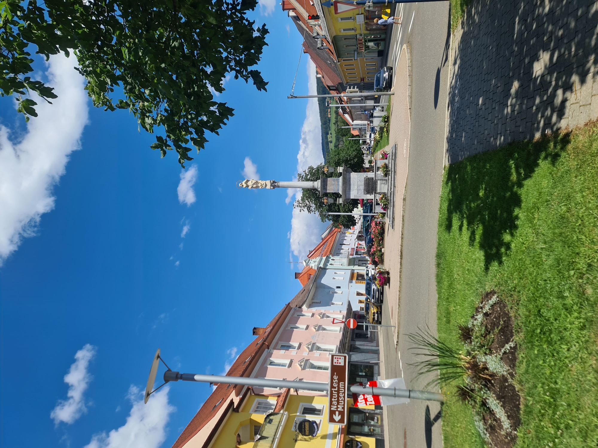 This is a street-level, full shot of a town square in Sankt Marein bei Neumarkt, Austria, on a clear, sunny day with scattered clouds. The perspective is from a sidewalk looking across a street intersection.
In the foreground to the left, there is a circular garden bed with green grass surrounding it, featuring some low-lying plants. To the right, a paved sidewalk runs parallel to the street, with dappled shade from trees overhead.
The midground is dominated by a cobblestone roundabout with a large monument in the center. The monument is a tall, stone column topped with a statue of a robed figure, possibly a religious icon. The base of the monument is surrounded by flower beds. Cars are visible driving on the road around the roundabout, and parked cars line the opposite side of the street.
The background features a row of colorful buildings lining the street, typical of Austrian town architecture. These buildings are of varying heights and colors, including yellow, pink, light blue, and green. Some have awnings and signs. Further in the distance, rolling green hills with scattered trees are visible under the bright blue sky. A street lamp is present in the left midground, and a traffic signal with a red light is visible on the right. A person in a blue dress is walking across a pedestrian crossing on the right side of the image.