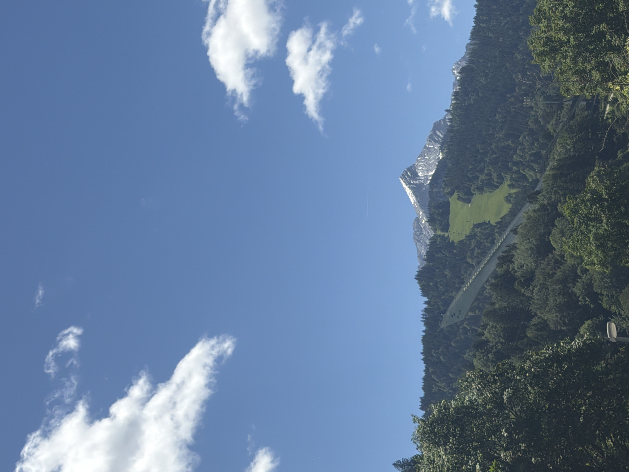 The image displays a majestic mountain peak under a clear blue sky dotted with white, fluffy clouds. The mountain is covered with a dense forest, and a vibrant green ski slope is visible on its side. The foreground is filled with lush green trees, and a street lamp can be seen in the lower left corner. The overall scene suggests a serene, natural landscape.