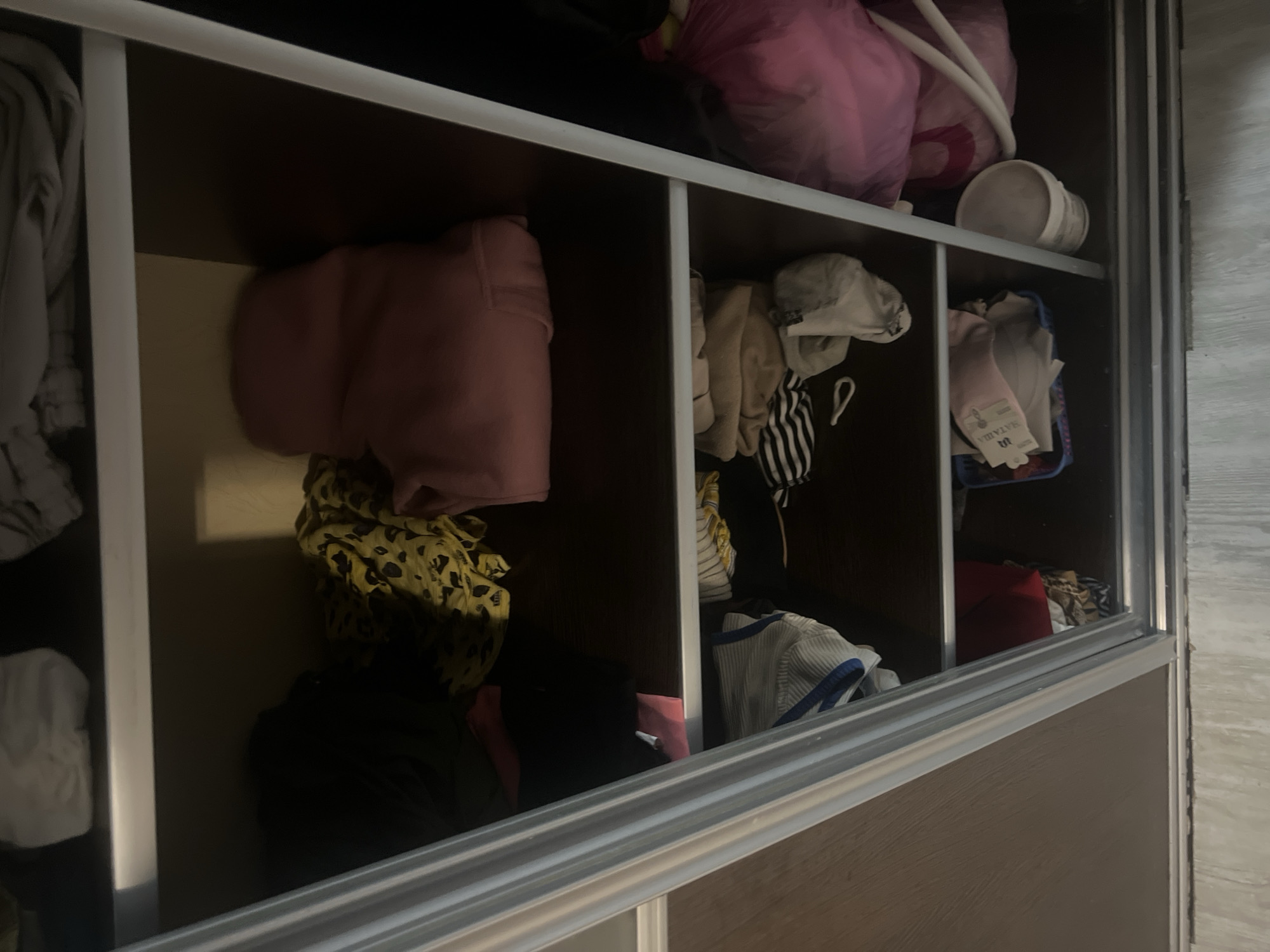 The image shows a cluttered closet with clothing and other items on shelves. The closet has sliding doors with a metallic frame. On the top shelf, there are folded items of clothing. The second shelf contains a black garment, a yellow leopard-print fabric, and a pink hooded sweatshirt. The third shelf has several items of clothing, including a striped garment and some folded beige fabric. On the bottom shelf, there is a blue basket containing folded items, a white bucket, and a large pink plastic bag. The floor outside the closet appears to be made of wood paneling. The location is described as Veteran-2, Russia, but no specific visual cues in the image confirm this location.