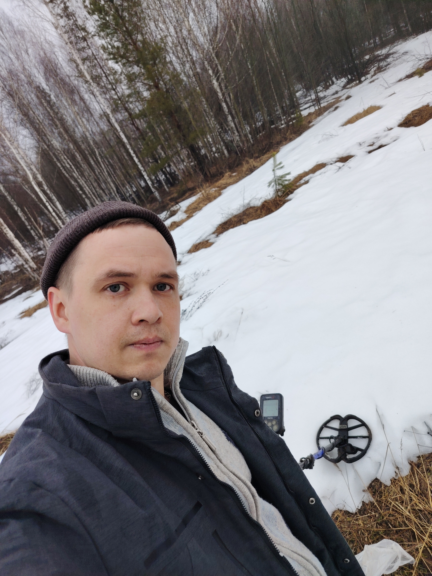 A selfie depicts a man in Марковский, Russia in a snowy, wooded area. He wears a dark gray beanie, gray sweater, and dark gray jacket. He holds a metal detector, whose screen and coil are visible in the foreground snow. The background shows a slope partially covered in snow, with dry grass patches and bare trees.