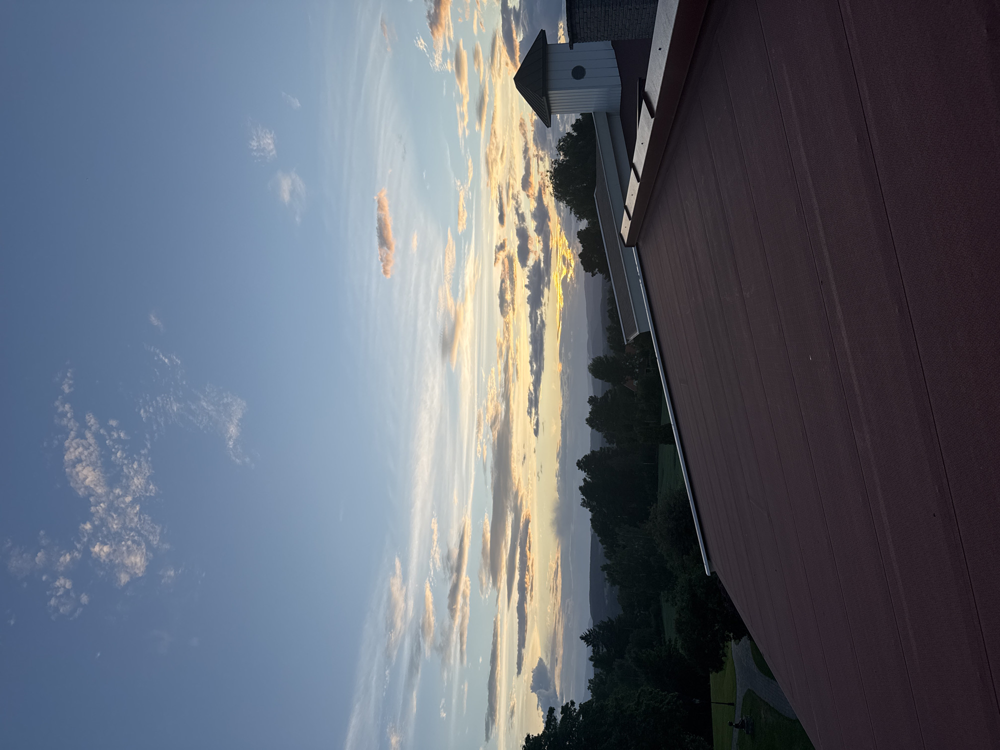 The image captures a wide-angle view from a rooftop in Hønefoss, Norway, during what appears to be sunset or twilight. The sky is a canvas of blues and soft oranges, with scattered clouds illuminated by the setting sun. In the foreground, a dark reddish-brown roof dominates the lower portion of the frame. To the right, a white, octagonal structure with a dark conical roof, resembling a small tower or ventilation shaft, is visible on the rooftop. Beyond the rooftop, a dense line of dark green trees stretches across the middle ground, with rolling hills fading into the distance under the colorful sky. The overall scene suggests a peaceful, natural landscape viewed from an elevated urban or suburban vantage point.