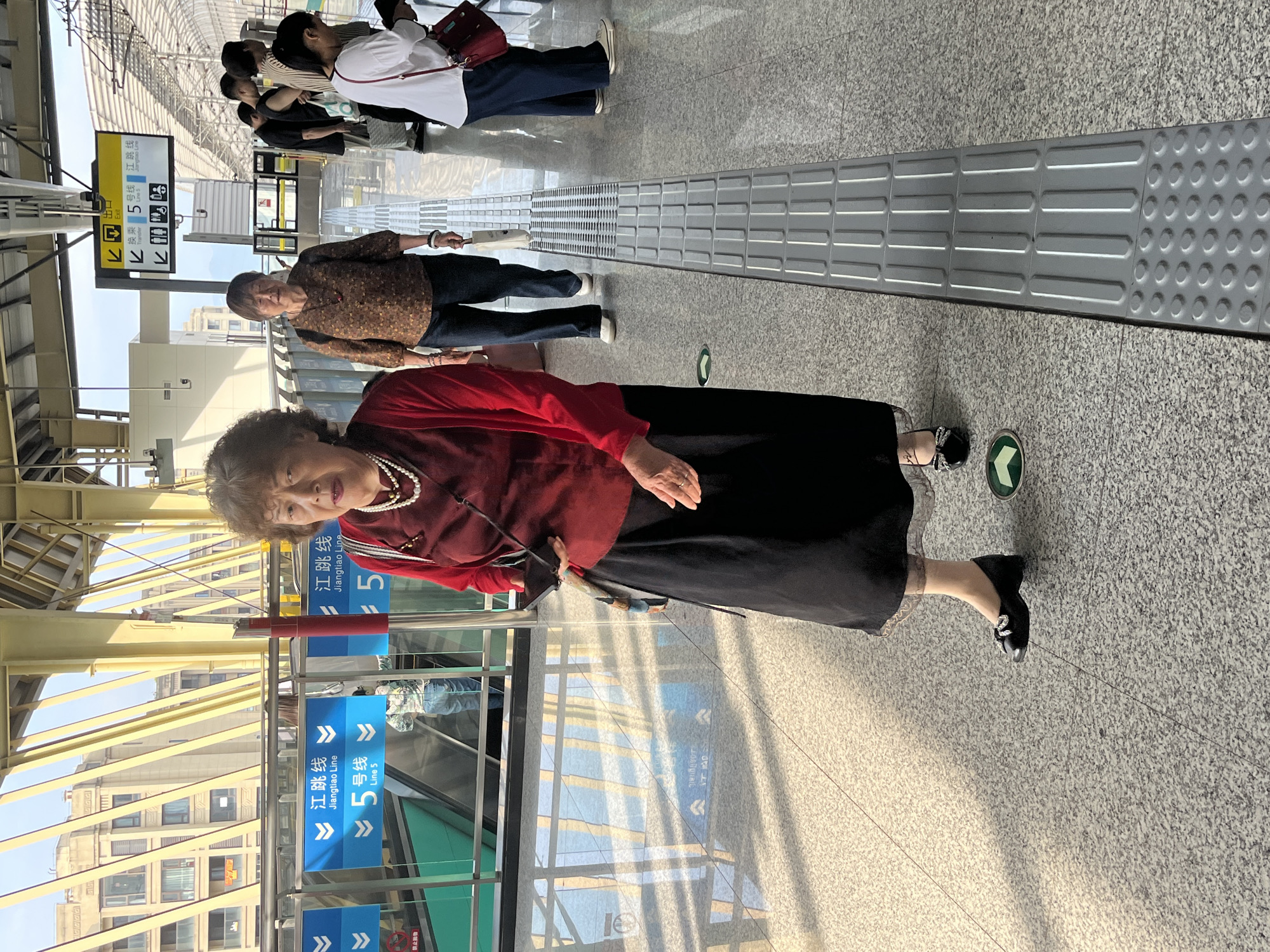 The image depicts an indoor scene at what appears to be a subway or train station platform in Tiaodeng, China. In the foreground, an elderly woman with dark, curly hair and wearing a red cardigan over a black skirt and black shoes with a bow detail is standing and looking towards the camera. She has a pearl necklace and is holding a dark object, possibly a phone, in her left hand. Behind her and to her right, another elderly woman in a brown patterned blouse and dark pants is walking away from the camera. Several other individuals, including a woman in a white top and dark pants with a red handbag, are visible in the background, along with signage indicating "Jiangtiao Line" and "Line 5." The platform floor is tiled with a grey, speckled pattern, and a tactile paving strip runs along the edge. Sunlight streams in from the left, casting shadows on the floor. The environment includes modern architectural elements, such as glass barriers and a metal ceiling structure.