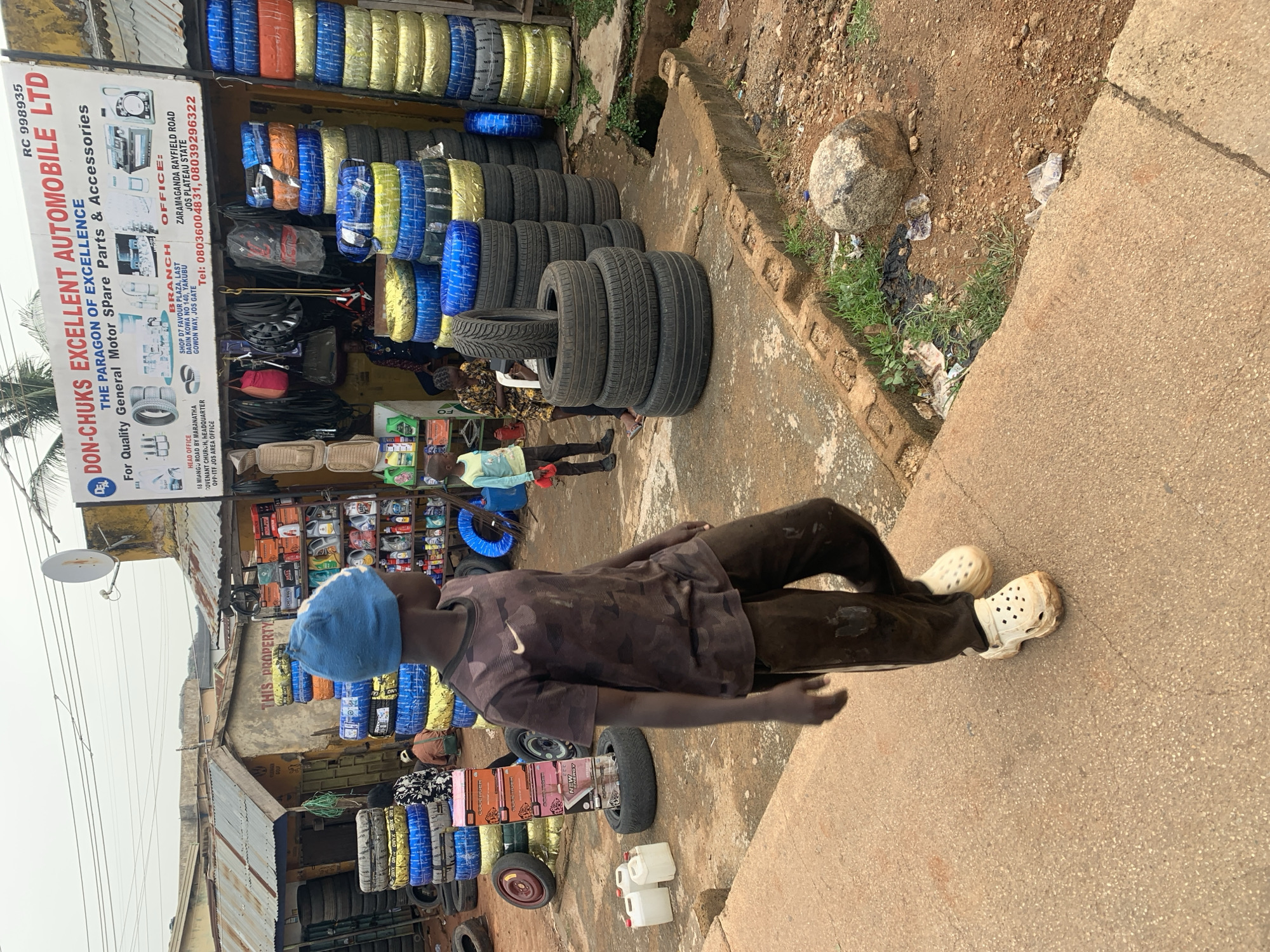 In Jos, Nigeria, a young boy wearing a blue beanie and a camouflage t-shirt walks past a tire shop. The shop, "DON-CHUKs EXCELLENT AUTOMOBILE LTD," displays stacks of new tires wrapped in blue, yellow, and black plastic. Various car parts and accessories are visible on shelves behind the counter. Two other children are also present near the shop, one holding a red object. The ground is a mix of dirt and concrete, with scattered debris. A large boulder sits on the side of the road. The scene depicts a busy street with commercial activity.