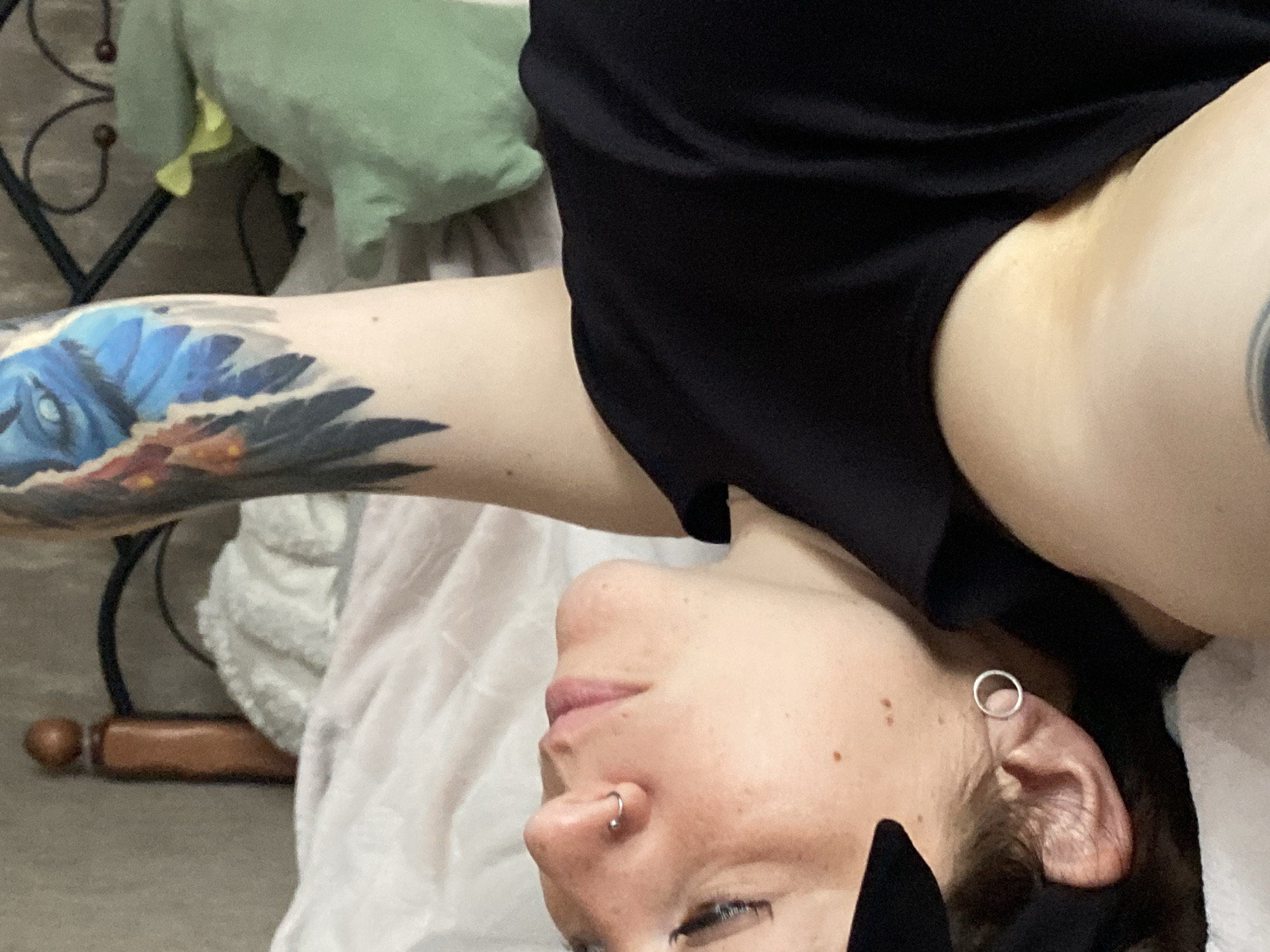A person is lying on a bed, their body oriented diagonally in the frame. The person has fair skin, visible freckles, and is wearing a black t-shirt. They have a hoop earring in their left ear and a nose ring. A large, colorful tattoo of what appears to be a bird's face with blue and orange feathers covers their left arm. The background includes a wooden bed frame, a white blanket, and a green stuffed frog. The scene is indoors, likely a bedroom. The location is Chaikovsky, Russia.