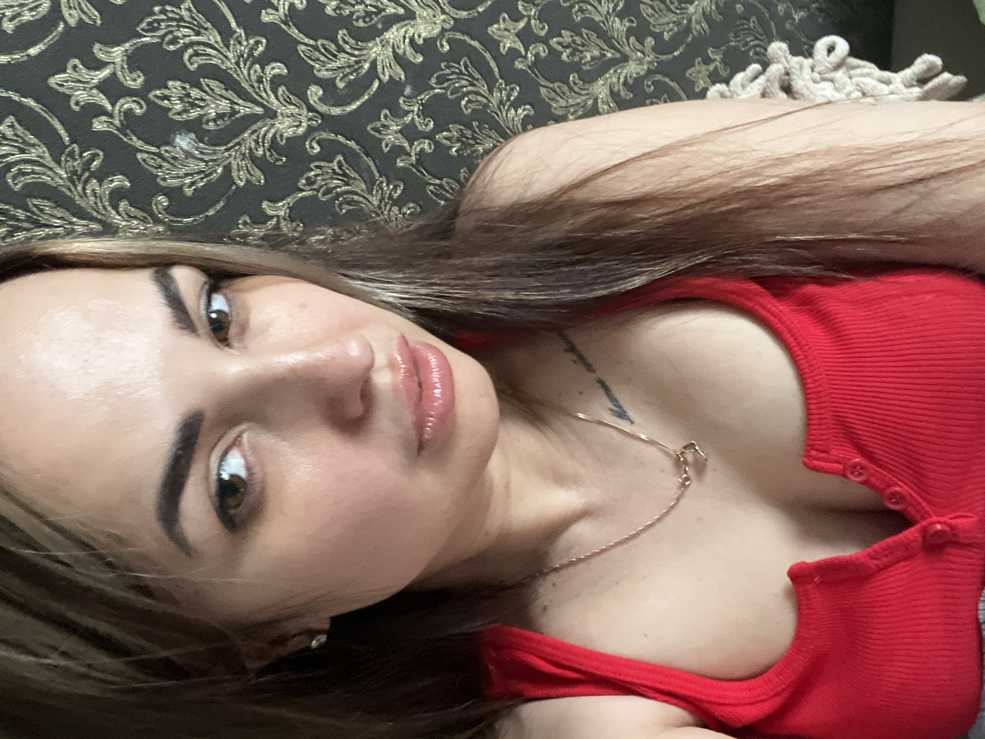 A female subject, appearing to be an adult, is depicted in a close-up, chest-up portrait, looking directly into the camera. She has long, multi-toned hair, predominantly brown with lighter streaks, falling over her shoulders. Her eyes are brown, and her eyebrows are dark and well-defined. Her lips are full and appear glossy or moist, slightly parted. A small, silver-colored stud earring is visible in her left earlobe. She is wearing a red, ribbed tank top or camisole with a partial front placket featuring three small buttons, the lowest of which is undone. A thin, gold-colored chain necklace with a small, indistinct pendant hangs around her neck. A script-style tattoo is partially visible on her left upper chest/shoulder area. The background consists of a dark-colored wall covered in wallpaper featuring an ornate, metallic gold, damask-like or floral scrollwork pattern. A small white smudge is visible on the wallpaper behind her right shoulder. A light-colored, textured object is partially visible on the far right side of the frame.