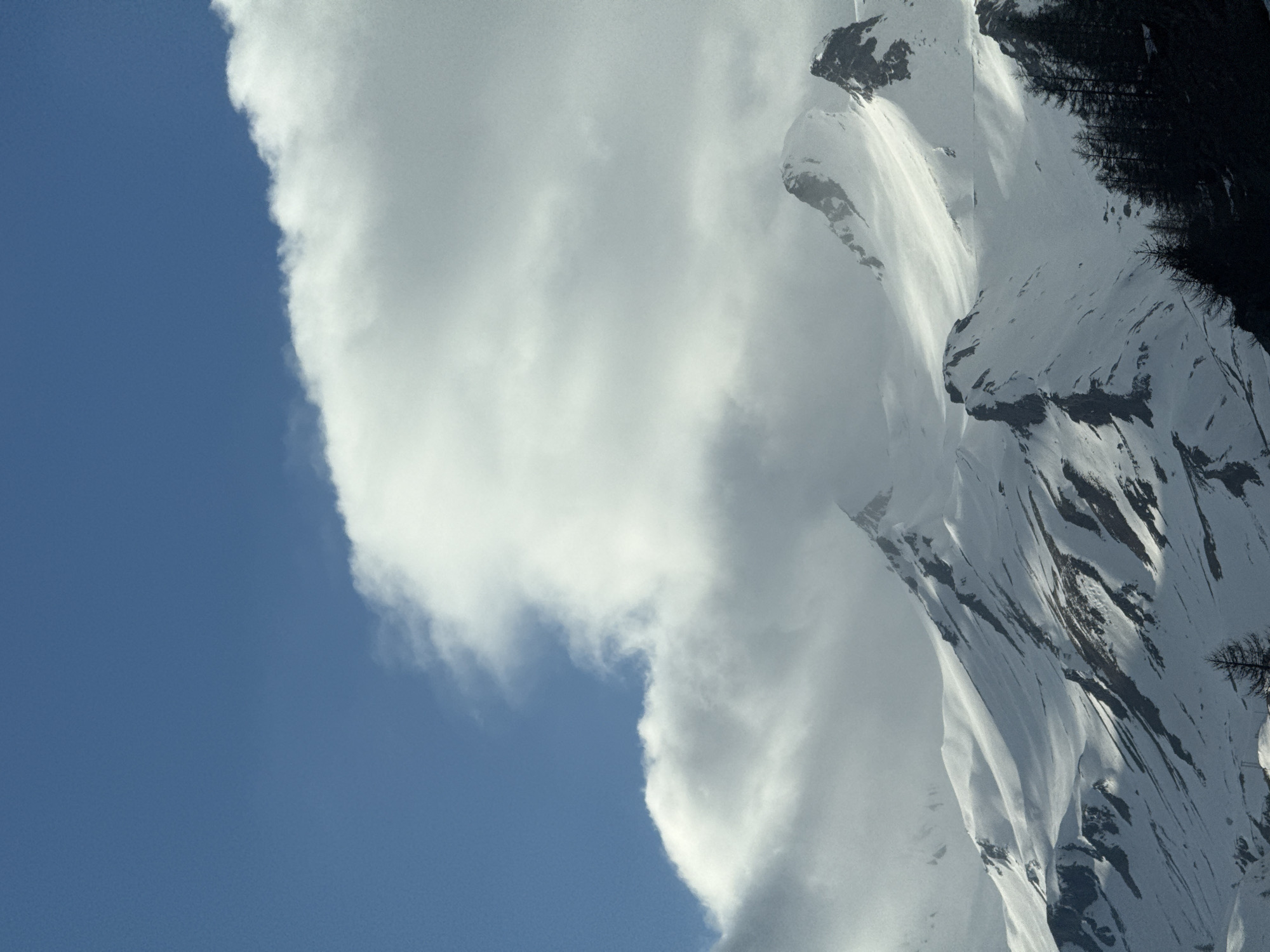 The image shows a partially snow-covered mountain range adjacent to a large cumulus cloud formation. A clear, blue sky is visible on the left side of the image. Dark green trees are present on the upper right side of the frame, along the mountain's slope. The scene appears to be photographed in daylight. The location is identified as Le Monêtier-les-Bains, France.