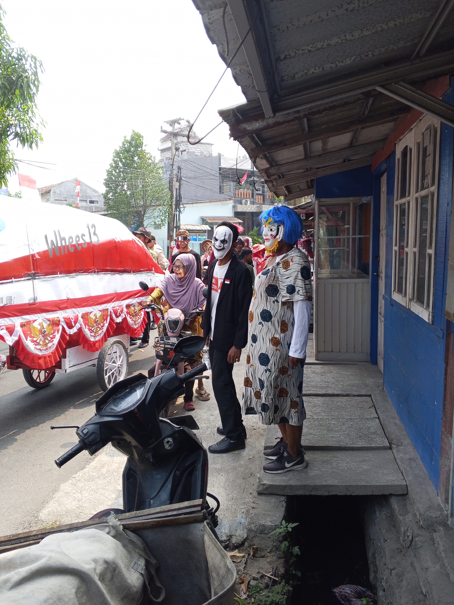 The image depicts a street scene in Ciruas, Indonesia, during what appears to be a local parade or festival. In the foreground, a black scooter is parked, partially obscuring a large, canvas-covered cart adorned with red and white banners featuring the Indonesian national emblem. The cart is also decorated with a fringe of red and white fabric. Behind the cart, several people are visible. A woman wearing a purple hijab and glasses rides a pink scooter. To her right, a person in a black suit and a white, skull-like mask stands next to another individual wearing a white patterned dress and a vibrant blue wig, also masked with a colorful, decorative mask. The background shows buildings, trees, and utility poles, consistent with an urban or semi-urban Indonesian setting. The overall atmosphere suggests a community event characterized by festive attire and masks.