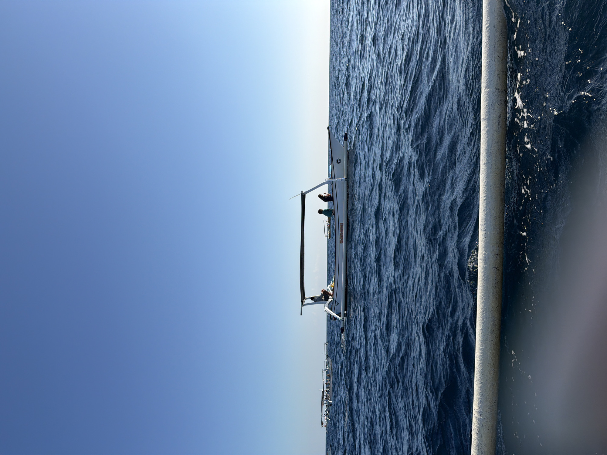 A wide shot captures a bright, clear day with a deep blue sky above a calm, dark blue ocean. Several white boats with outriggers are visible. In the foreground, a white tube or buoy runs horizontally across the bottom of the frame. The closest boat, in the mid-ground, is a motorboat with a canopy, carrying at least three people. One person is standing near the stern, appearing to steer, while two others are seated. Text in orange is visible on the side of this boat. Further in the distance, two other motorboats are also visible, each carrying multiple passengers. The scene suggests a leisurely outing on the water, likely near Seririt, Indonesia, given the style of the boats which are characteristic of traditional Indonesian fishing vessels adapted for tourism.