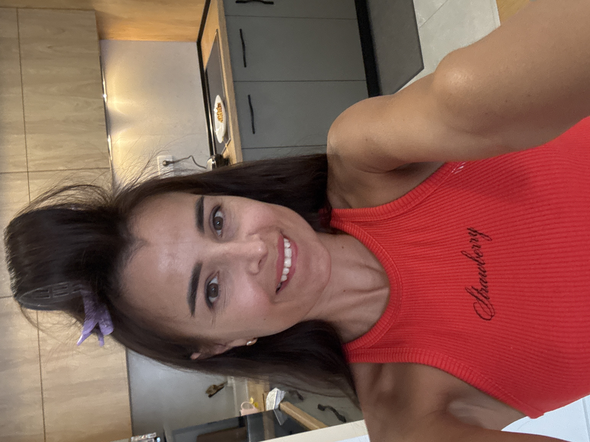 A woman with brown hair pulled up with a purple clip, wearing a red ribbed tank top with the word "Strawberry" embroidered on it, is taking a selfie in a modern kitchen. The kitchen has light wood cabinets above dark grey cabinets. A plate with food is on the stovetop. The woman is smiling and looking at the camera.