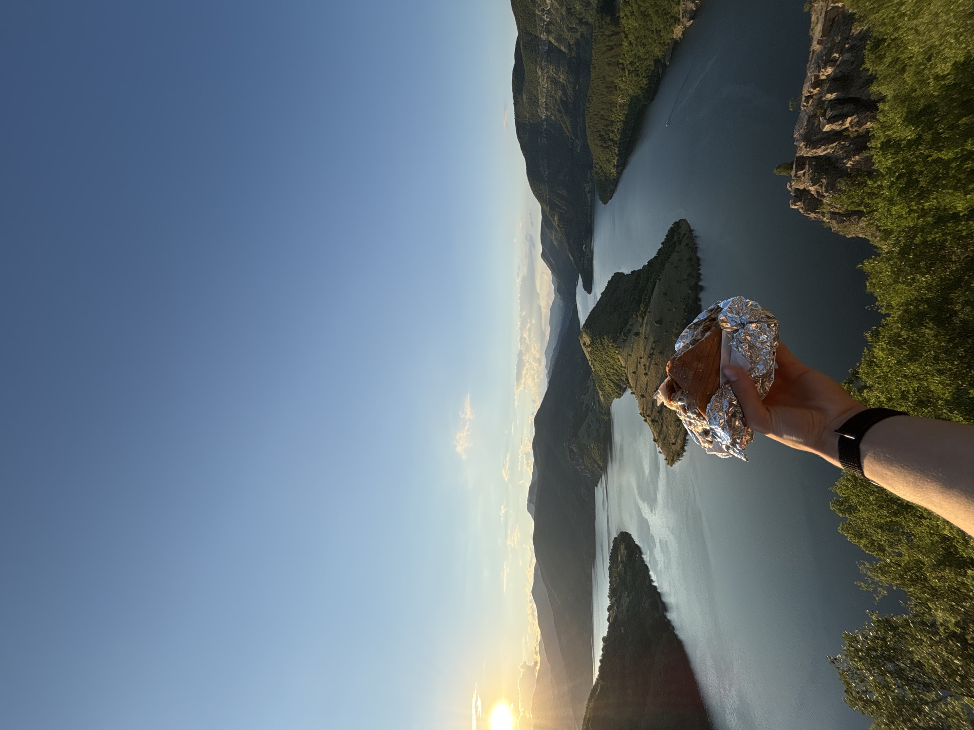 A person's arm and hand are visible in the foreground, holding a sandwich wrapped in aluminum foil. The sandwich is positioned in front of a scenic vista of a wide river or lake winding through lush, tree-covered mountains. The sun is setting or rising on the left side of the image, casting a warm glow and creating a silhouette of the mountains. The sky is a clear blue with a few scattered clouds. The location is identified as Borovitsa, Bulgaria.