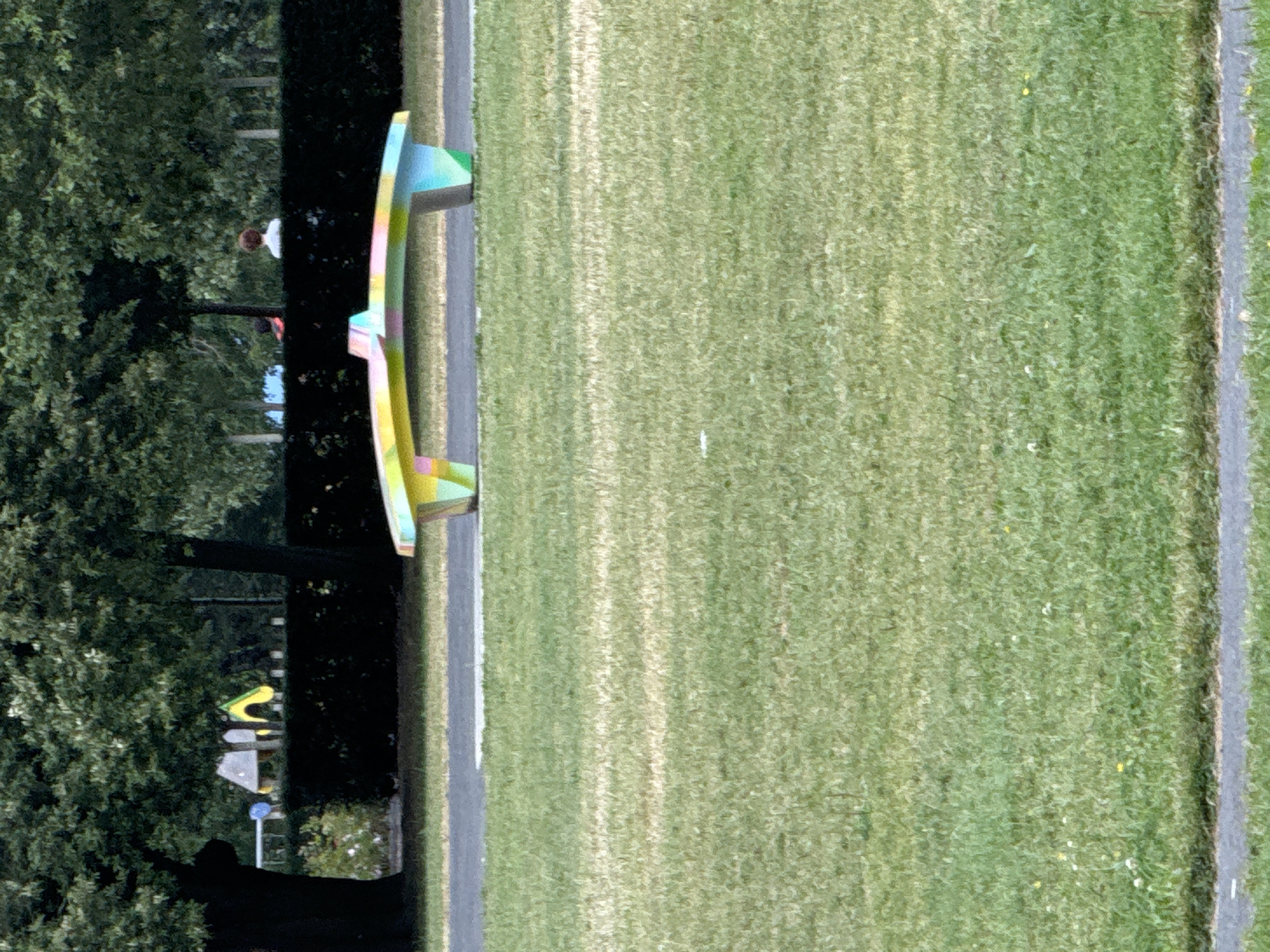This is a medium-range, eye-level, outdoor shot of a park on a bright day, possibly in Waterford, Ireland. The foreground is dominated by a wide expanse of well-maintained green grass, with a grey paved path running horizontally across the bottom of the frame. In the middle ground, a vibrantly colored, modern bench with a curved design, painted in a rainbow of pastels, sits on a grey paved area. Behind the bench, a dense dark green hedge runs across the image, forming a backdrop. Beyond the hedge, two people are visible: one with dark hair and a red shirt on the left, and another with curly brown hair and a white shirt on the right, standing near some trees. The background consists of more trees and some playground equipment, including a yellow slide and blue and grey structures. The overall impression is one of a tranquil public park.