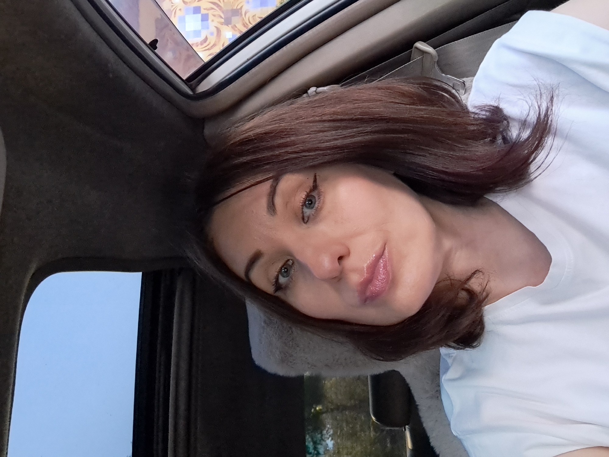 A woman with shoulder-length dark brown hair is taking a selfie inside a car. She is wearing a white t-shirt. Her makeup includes winged eyeliner and lip gloss. The car's sunroof is open, revealing a light blue sky. The interior of the car, including a beige seat and the dark gray headliner, is visible. The image does not provide any specific information about the location in Irkutsk, Russia.