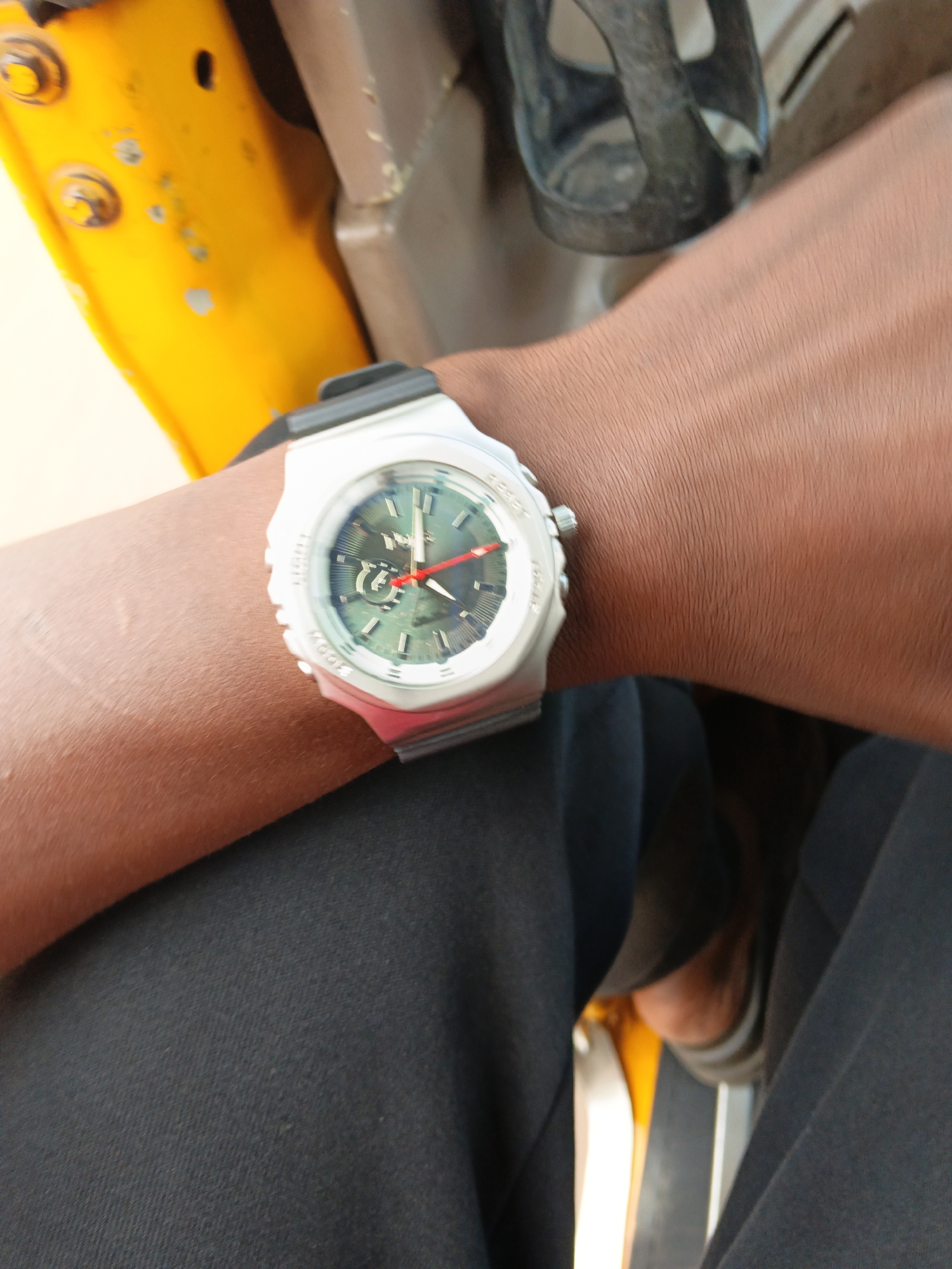 This media file is a close-up, eye-level shot of a person's arm wearing a watch. The person is wearing dark pants and is possibly sitting in or near a yellow vehicle, with parts of the vehicle visible in the background. The watch has a white case and a dark band, with a green analog dial and red hour and minute hands. There are also some markings on the bezel of the watch, possibly indicating functions like "LIGHT" and "MODE." The lighting suggests it is daytime, but no specific time or weather conditions are evident. The overall tone is neutral, focusing on the watch and the immediate surroundings.