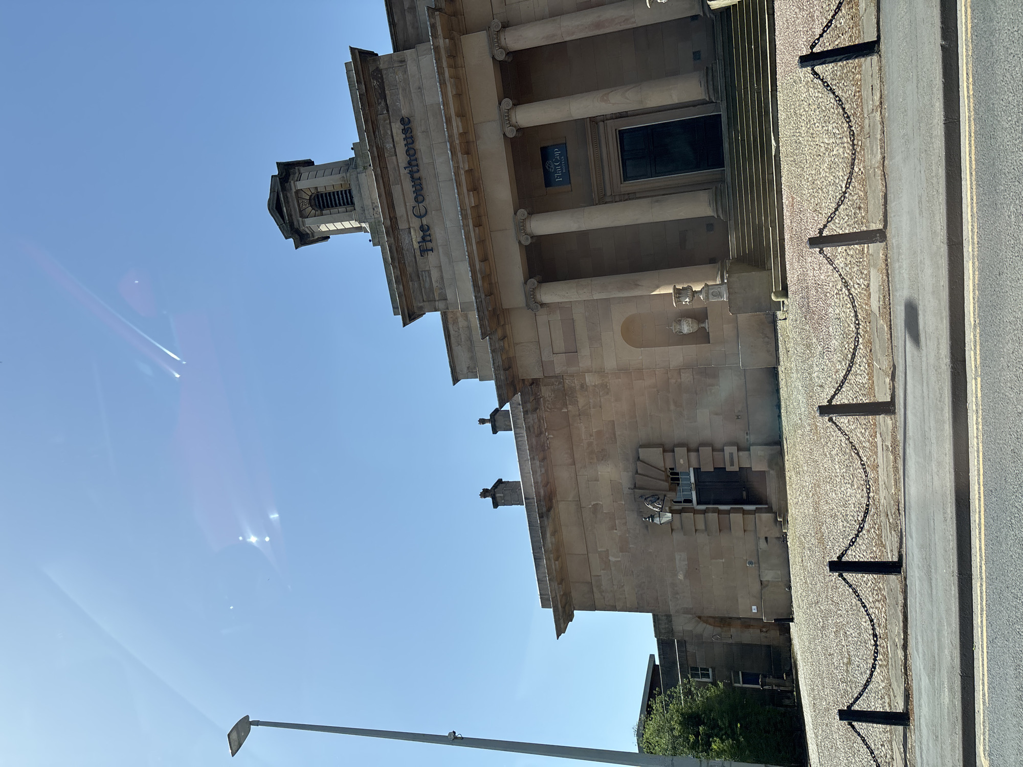 The image captures an upward angle of "The Courthouse" building in Knutsford, United Kingdom, under a clear blue sky. The building, made of light brown stone, features a classical architectural style with a prominent sign displaying "The Courthouse" and "Flat Cap Hotels". Its facade includes columns, a tower-like structure on top, and an ornate entrance. A chain fence runs along the perimeter, separating the building from the street. A streetlight is also visible.