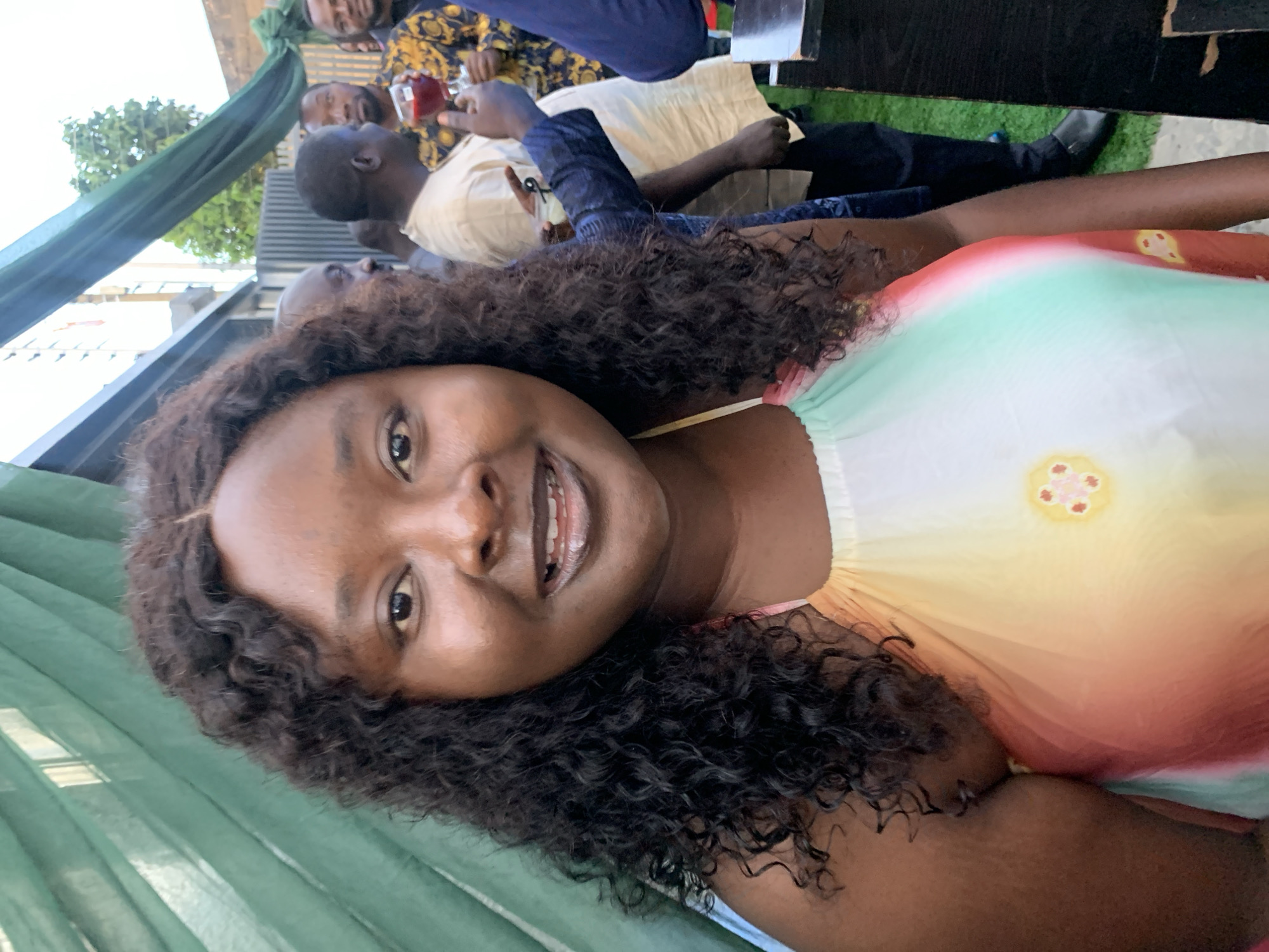 A woman with curly dark hair smiles at the camera in the foreground, wearing a halter top with a tie-dye pattern of pink, orange, yellow, and green, adorned with floral motifs. Her makeup includes dark lipstick. Behind her, several men are visible, some holding drinks and engaging in conversation. One man in the mid-ground wears a patterned gold and black shirt, while another to his left wears a cream-colored garment. The background suggests an outdoor event with green draping and foliage. The scene appears to be set in Gura Top, Nigeria, during what seems to be a social gathering or celebration.