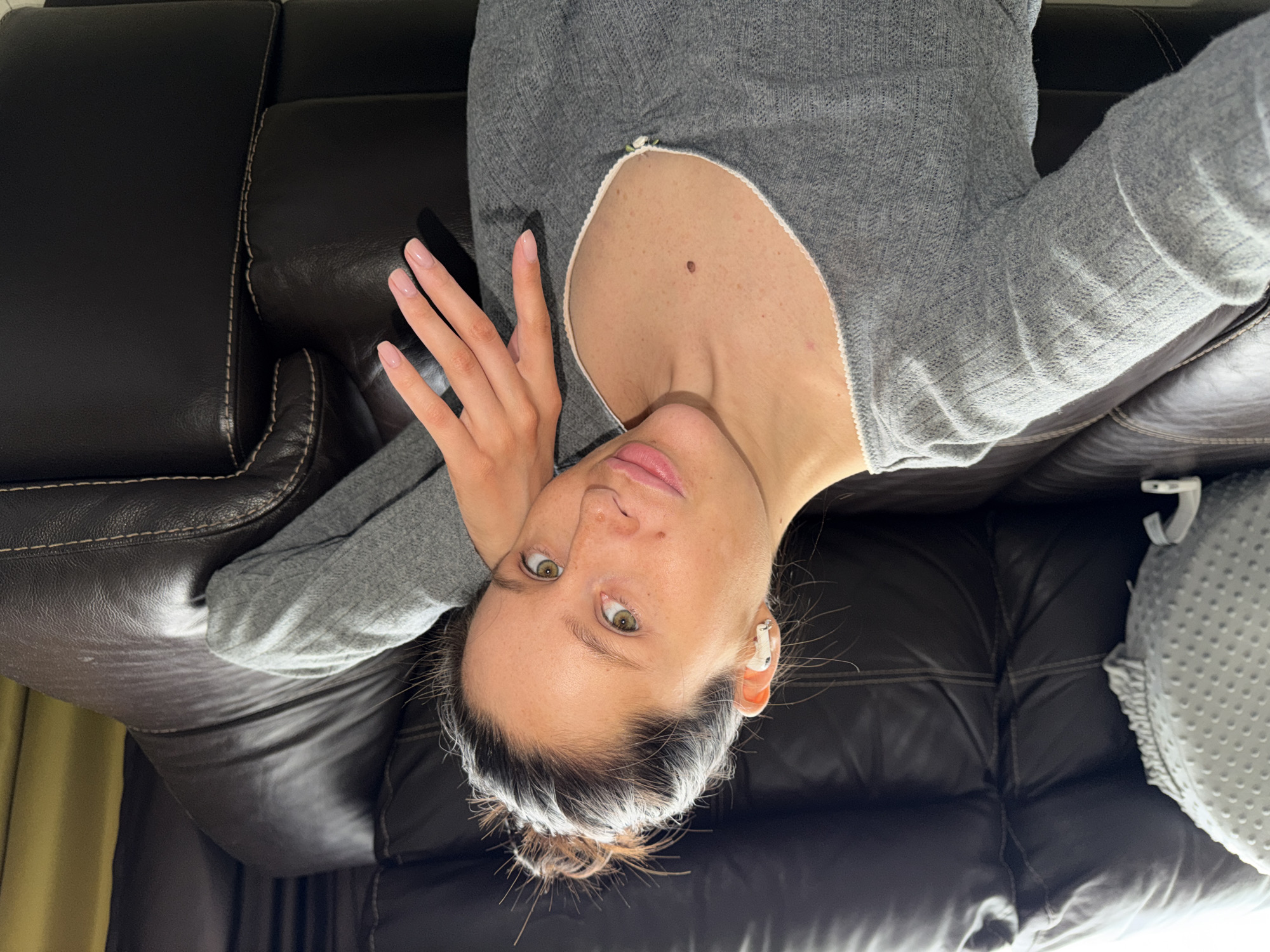 The image captures a close-up, eye-level selfie of a person, likely a woman, lying on a dark brown leather sofa. The setting is indoors, characteristic of a living room, and is specified as Khimki, Russia.
The subject has fair skin, striking light-colored eyes (appearing greenish-brown), and dark hair pulled back into a bun, with distinct grey or white strands visible at the hairline. She wears a relaxed, neutral expression, looking directly into the camera. Her attire consists of a soft, light grey, long-sleeved top with a wide, scooped neckline, revealing her upper chest where a prominent mole is visible.
Her right hand, adorned with neatly manicured nails painted in a subtle, natural pink or nude shade, rests near her shoulder. A white earbud is visible in her right ear. The dark brown leather sofa features visible stitching details, suggesting a comfortable and well-maintained piece of furniture. A light grey, textured pillow with a dotted pattern is partially visible on the right side of the frame, next to the subject.
The overall impression is one of a casual and relaxed moment, likely a self-portrait taken in a home environment. The lighting is soft and even, suggesting either diffused natural light or ambient indoor illumination. There is no visible text within the image.