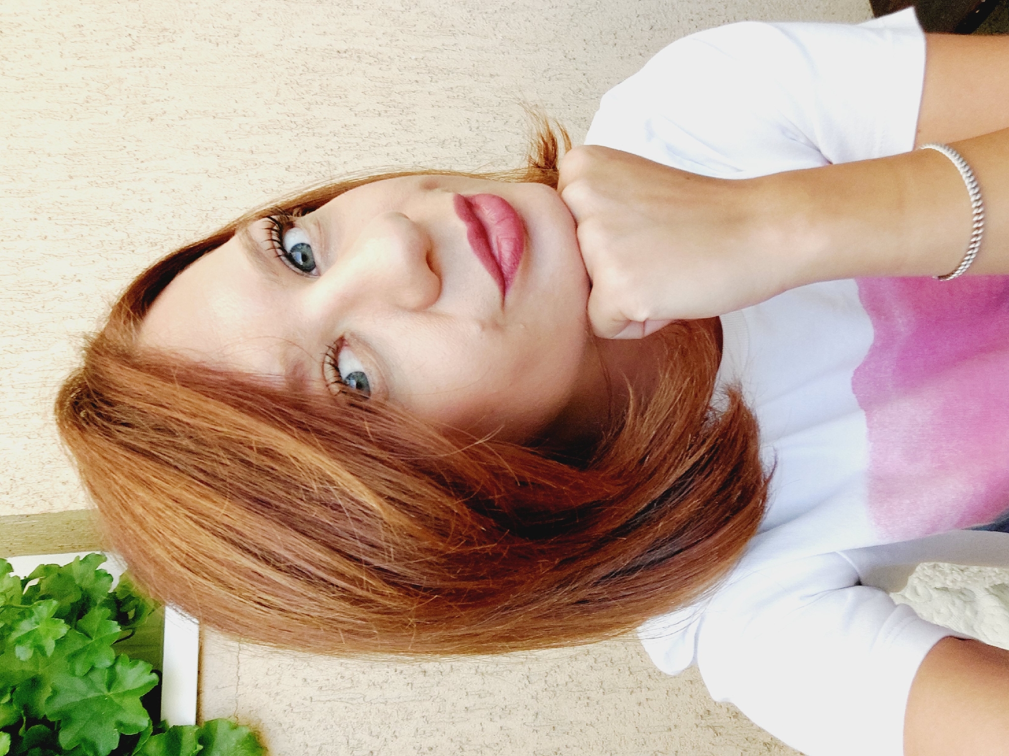 In Sofia, Bulgaria, a close-up selfie shows a woman with auburn hair and blue eyes, resting her chin on her hand. She is wearing a white t-shirt with a pink design and a silver bracelet. The background features a textured, light-colored wall and a hint of green foliage from a plant in the upper left corner.