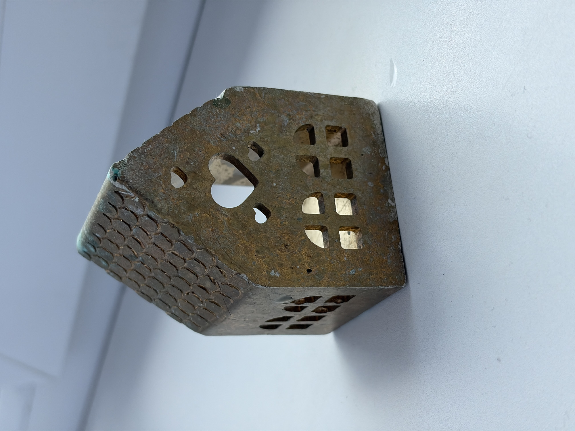 This is a close-up, eye-level, medium shot of a small, decorative metal house, possibly a candle holder or ornament, positioned against a white, textured wall. The house is made of a brass-like metal and has a worn, aged patina with some green oxidation visible. Its design features a patterned roof, possibly representing shingles, and cut-out windows in various shapes, including squares and a prominent heart shape on one side. The house is angled, with its left side facing the camera and a portion of its base visible. The background is a plain white wall with subtle texture and a few minor imperfections, suggesting an indoor setting. There are no people or other objects in the frame. The lighting is soft and diffuse, casting subtle shadows that highlight the textured surfaces. The overall mood is quiet and still. There is no visible text in the image.