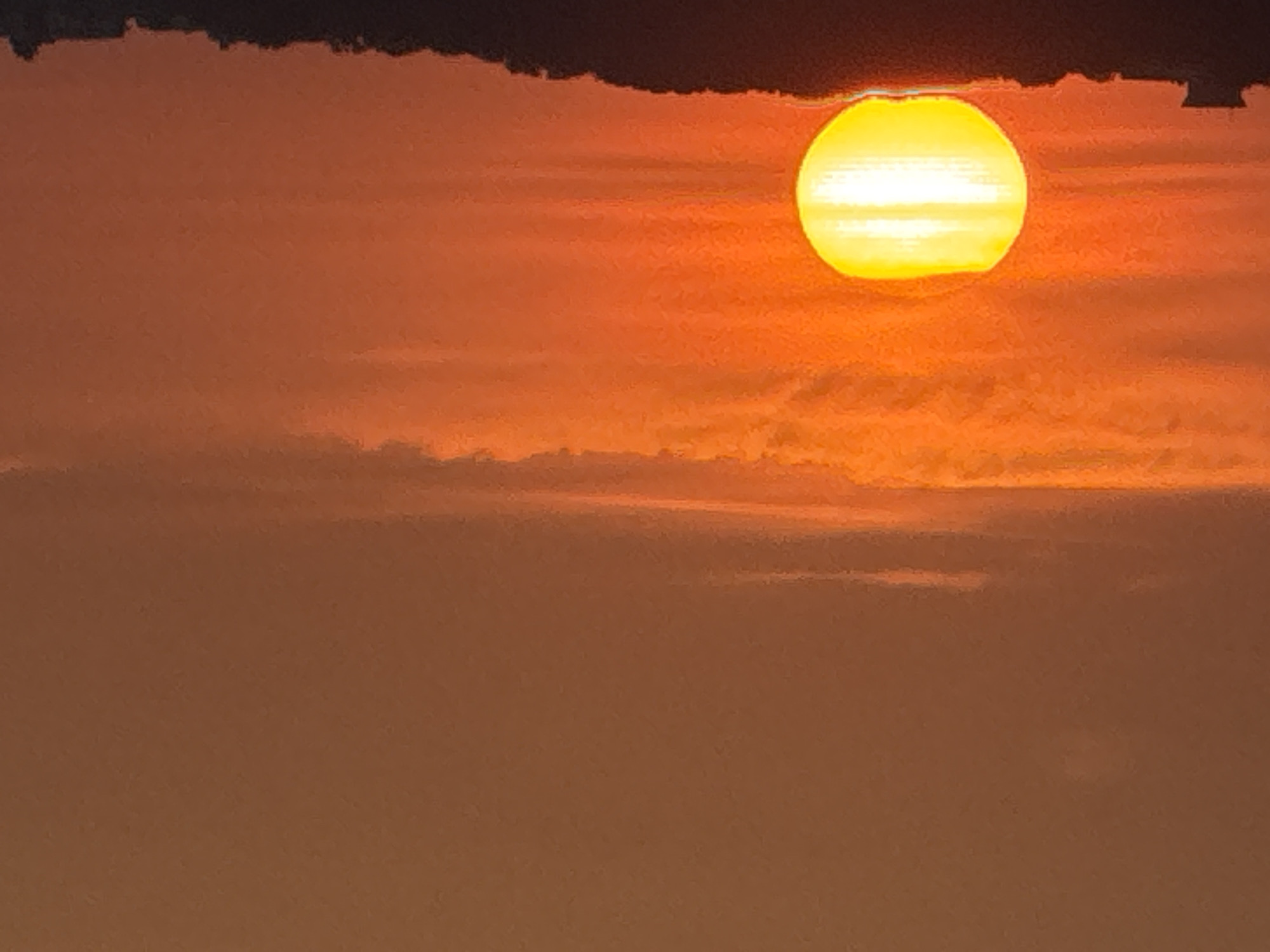 The image depicts a sunset over a silhouetted landscape of rolling hills or a treeline, with a building visible on the left side. The sun, a bright yellow disc with horizontal bands of light, is low in the sky, partially obscured by the horizon. The sky is a gradient of orange and reddish hues, with thin, horizontal clouds. The foreground landscape is dark and indistinct, suggesting it is either night or heavily shadowed. The location is specified as Unknown, Unknown.