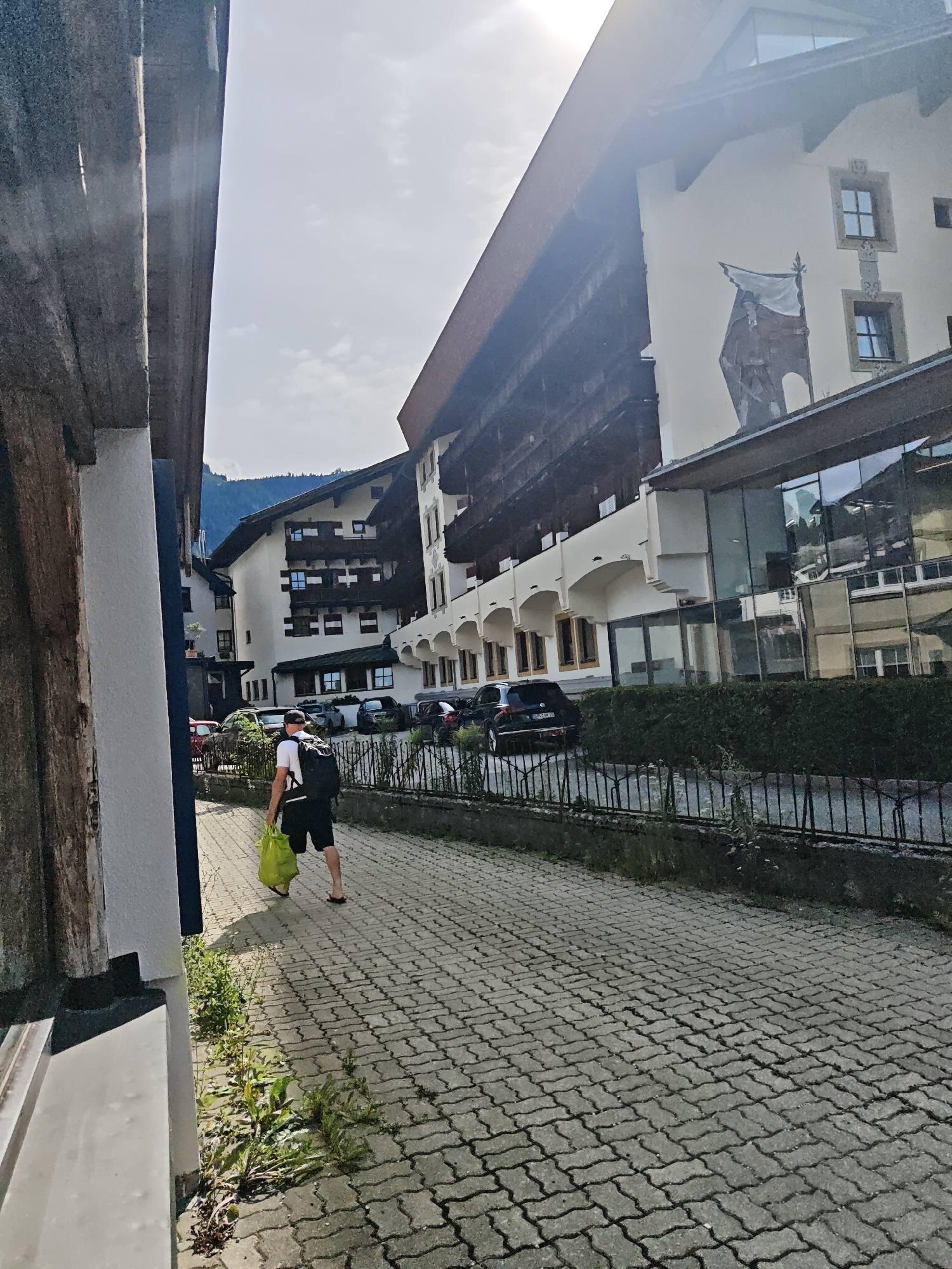 A person is walking on a cobblestone street in Kitzbühel, Austria, carrying a bright green plastic bag. The person is wearing a white t-shirt, black shorts, a black backpack, and flip-flops. They are walking away from the camera, towards a street lined with buildings and parked cars. The buildings are typical of Alpine architecture, with wooden balconies and ornate facades. One building features a large mural of a man holding a flag. The sky is bright and sunny, with some clouds visible. The street appears to be in a town or village setting.