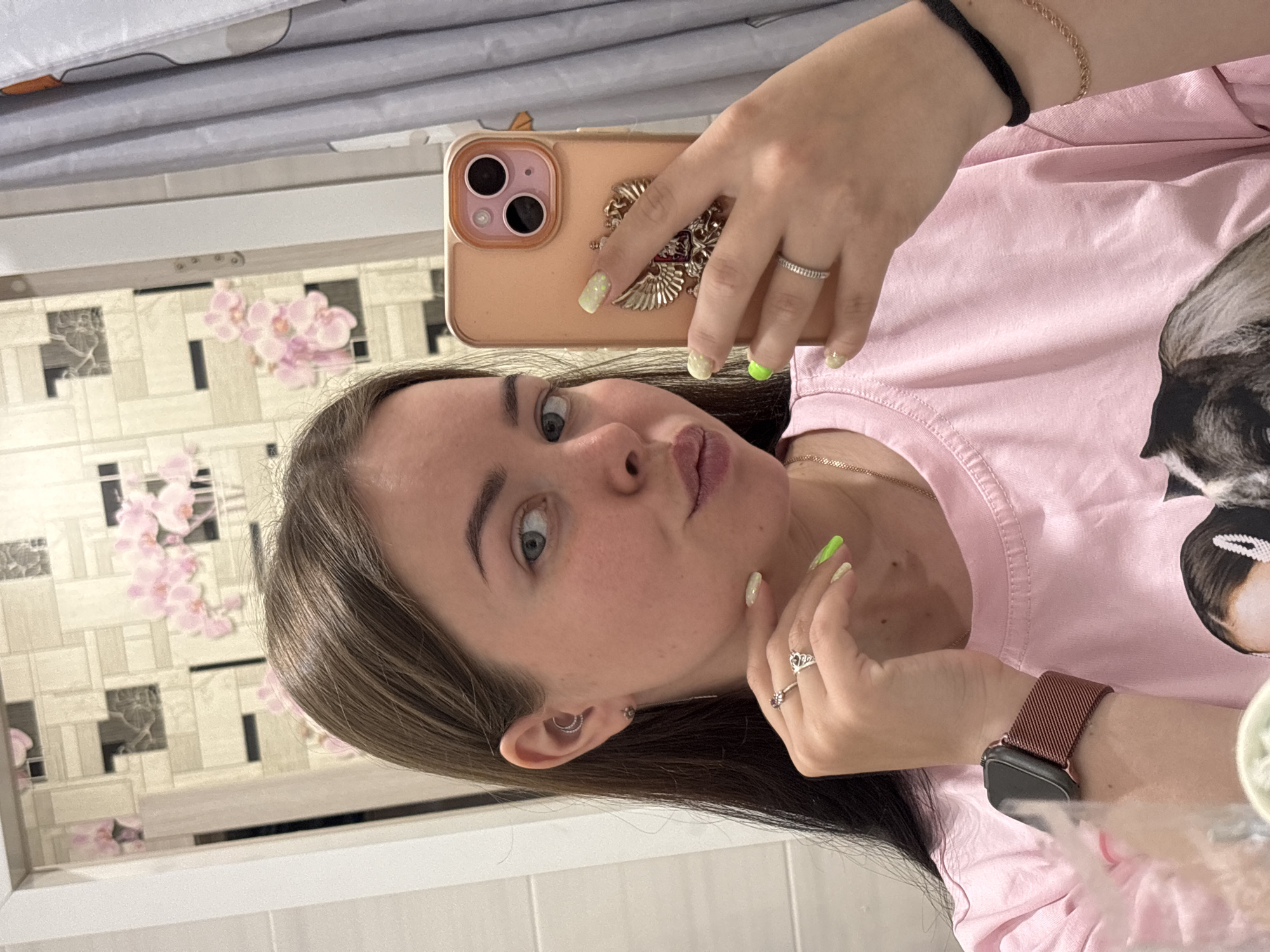 A young woman is taking a selfie in a bathroom. She is holding a peach-colored iPhone in her right hand, which has a decorative case. Her nails are painted in shades of yellow and green. She wears several rings on her fingers and an Apple Watch on her wrist. The woman is wearing a pink t-shirt. In the background is a tiled wall with a floral pattern. The location is potentially in Izhevsk, Russia.