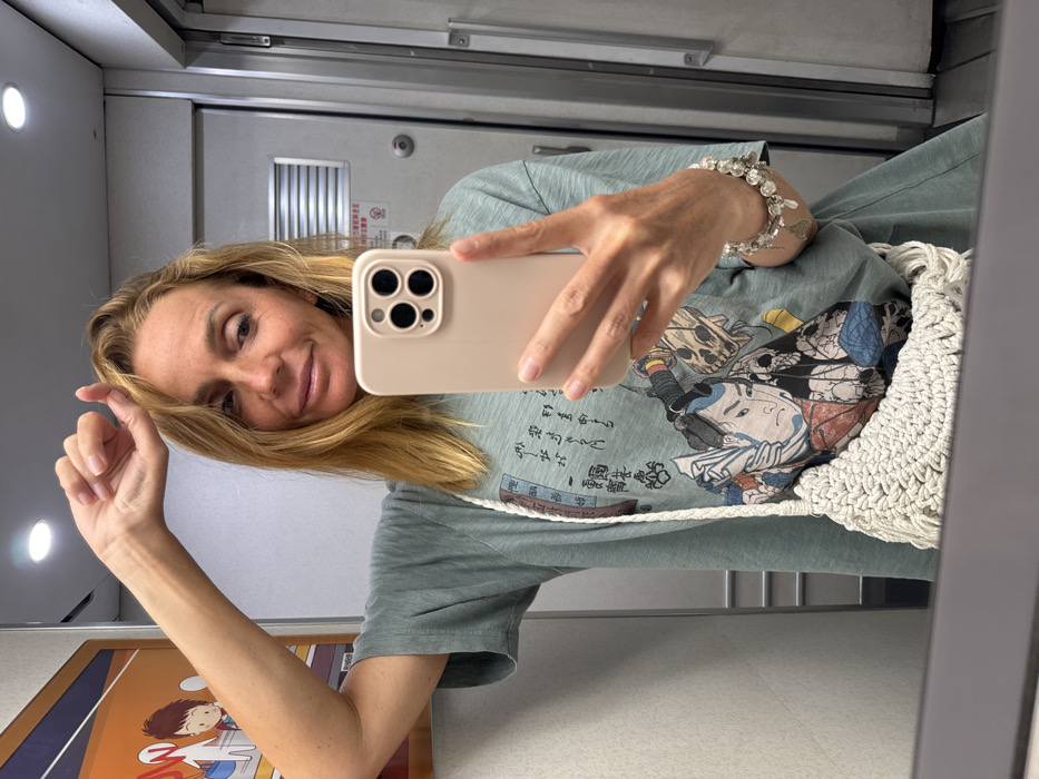A female subject is depicted taking a mirror selfie with a light-colored smartphone. The scene is set in the city of Dong'an, China, within what appears to be a train cabin or restroom. The subject, with shoulder-length blonde hair, gazes slightly to her right, a subtle smile on her lips. Her right arm is raised, with the hand partially obscuring the top of her head. She wears a light green oversized t-shirt featuring a Japanese ukiyo-e style print of a male figure with an elaborate headdress and a partial skull design, along with Japanese text. A white, crocheted shoulder bag with fringe hangs across her body. Her left wrist displays a multi-strand bracelet adorned with clear and silver-toned charms. The smartphone, held by her left hand, has a light-colored case and a prominent triple-lens camera array on its back. The background shows reflective surfaces, suggesting a mirror, with an overhead recessed light fixture. Beyond the reflection, elements of a train interior are visible: a light-colored door with a horizontal ventilation grate and a circular lock mechanism, and to the left, a framed cartoon poster depicting two stylized figures on an orange background.